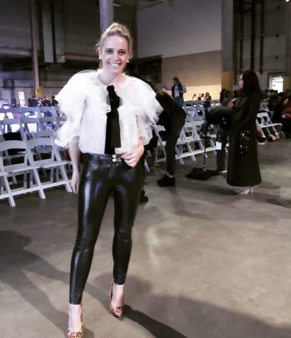 Woman in shiny black pants and white top standing in an indoor event space with chairs and people in background.