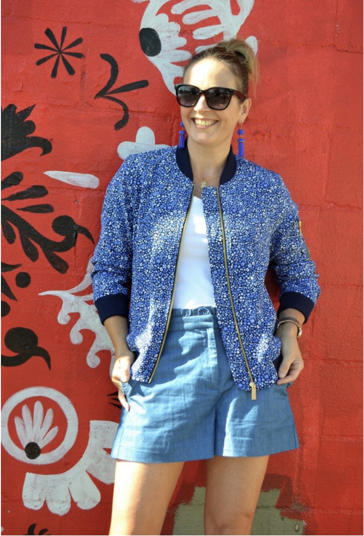Woman wearing sunglasses, blue jacket, and denim shorts standing in front of a red mural with floral and abstract designs.