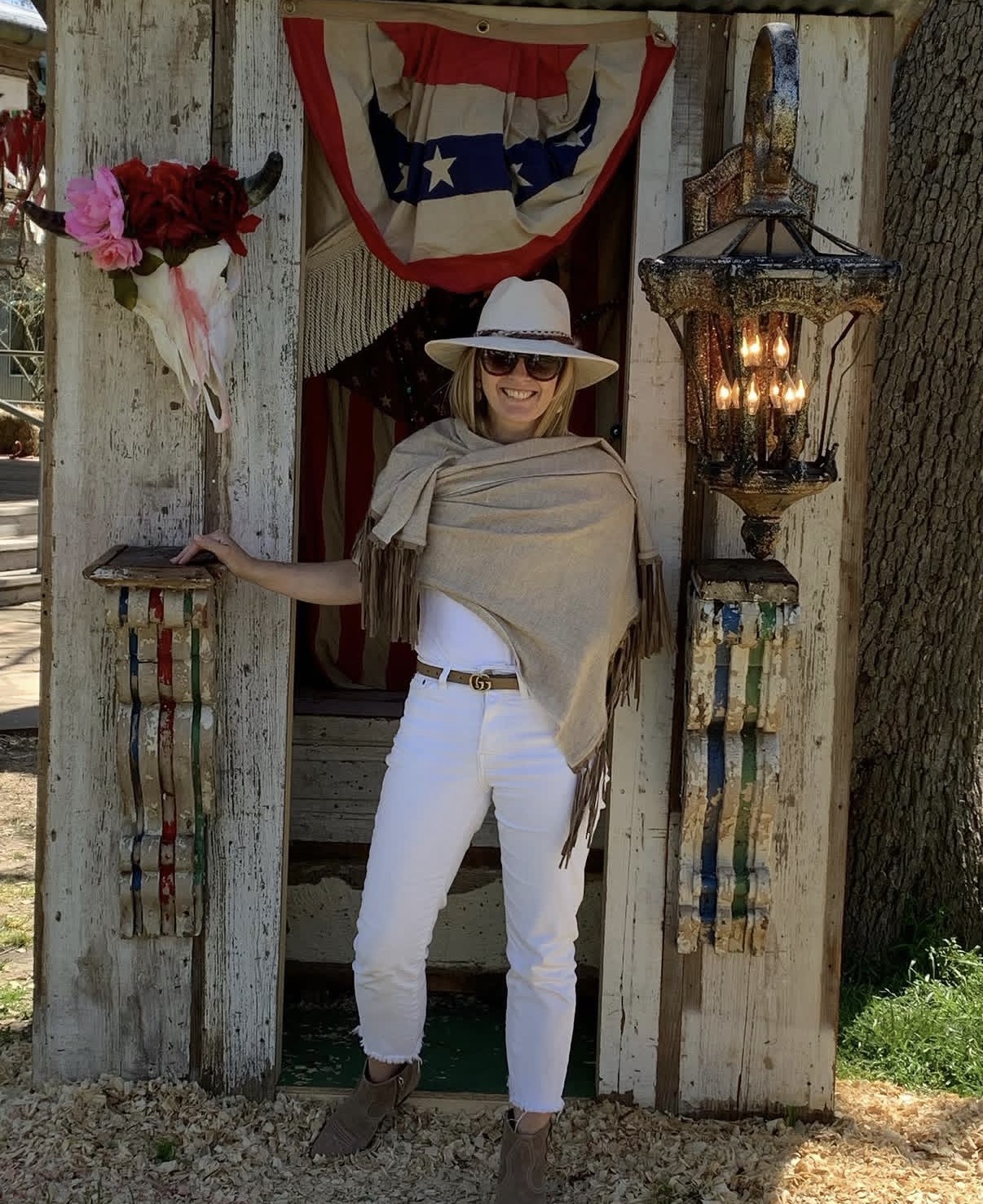 Woman wearing a wide-brimmed hat, sunglasses, beige shawl, and white pants standing in front of a rustic wooden structure with patriotic decorations.