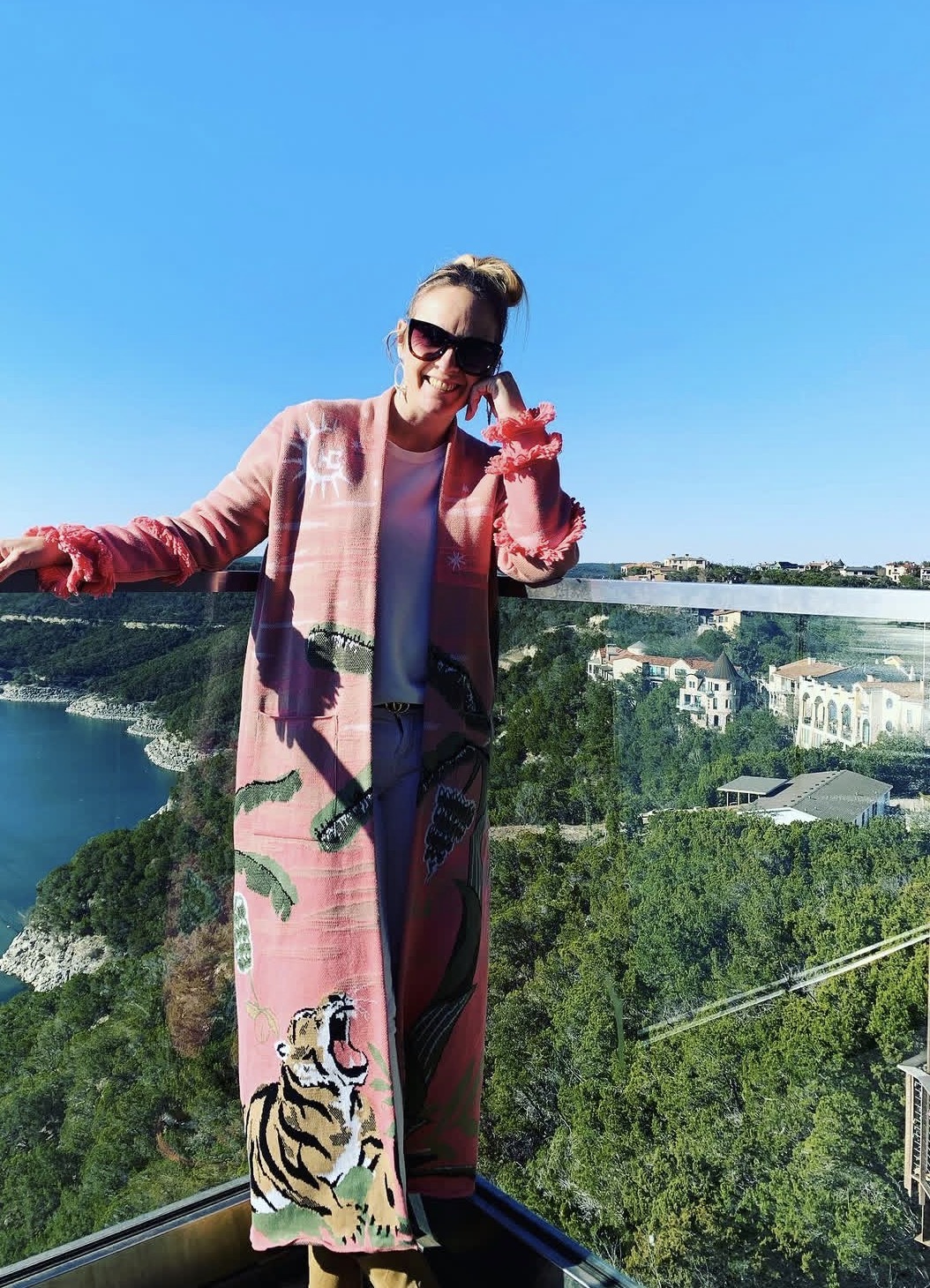 Woman in sunglasses and pink coat standing outdoors with a scenic view of trees and buildings behind her.