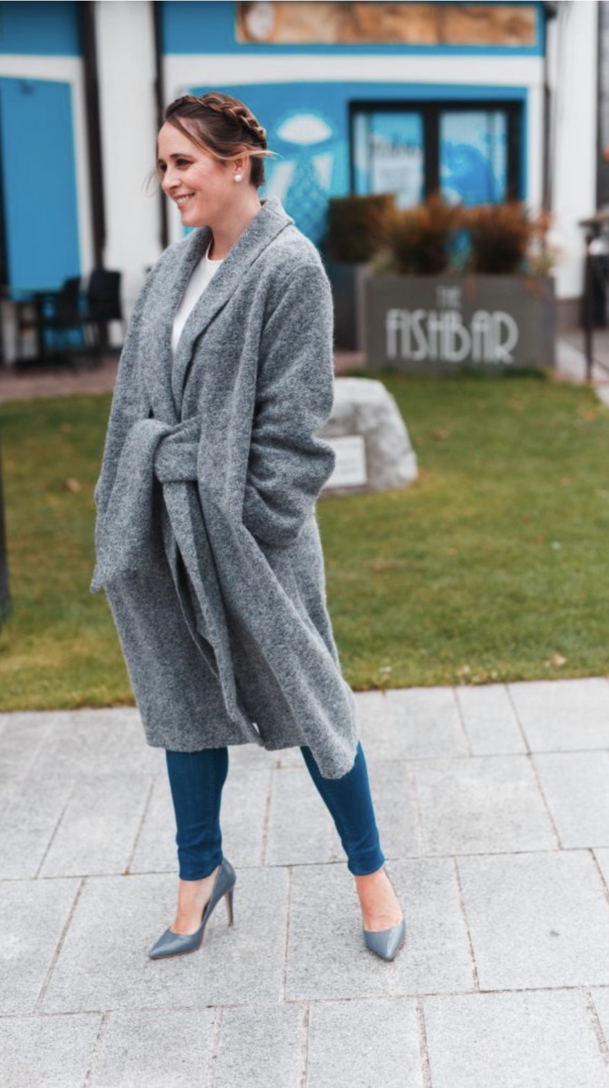 Woman in gray coat and high heels standing outdoors, smiling, with a building and sign in the background.