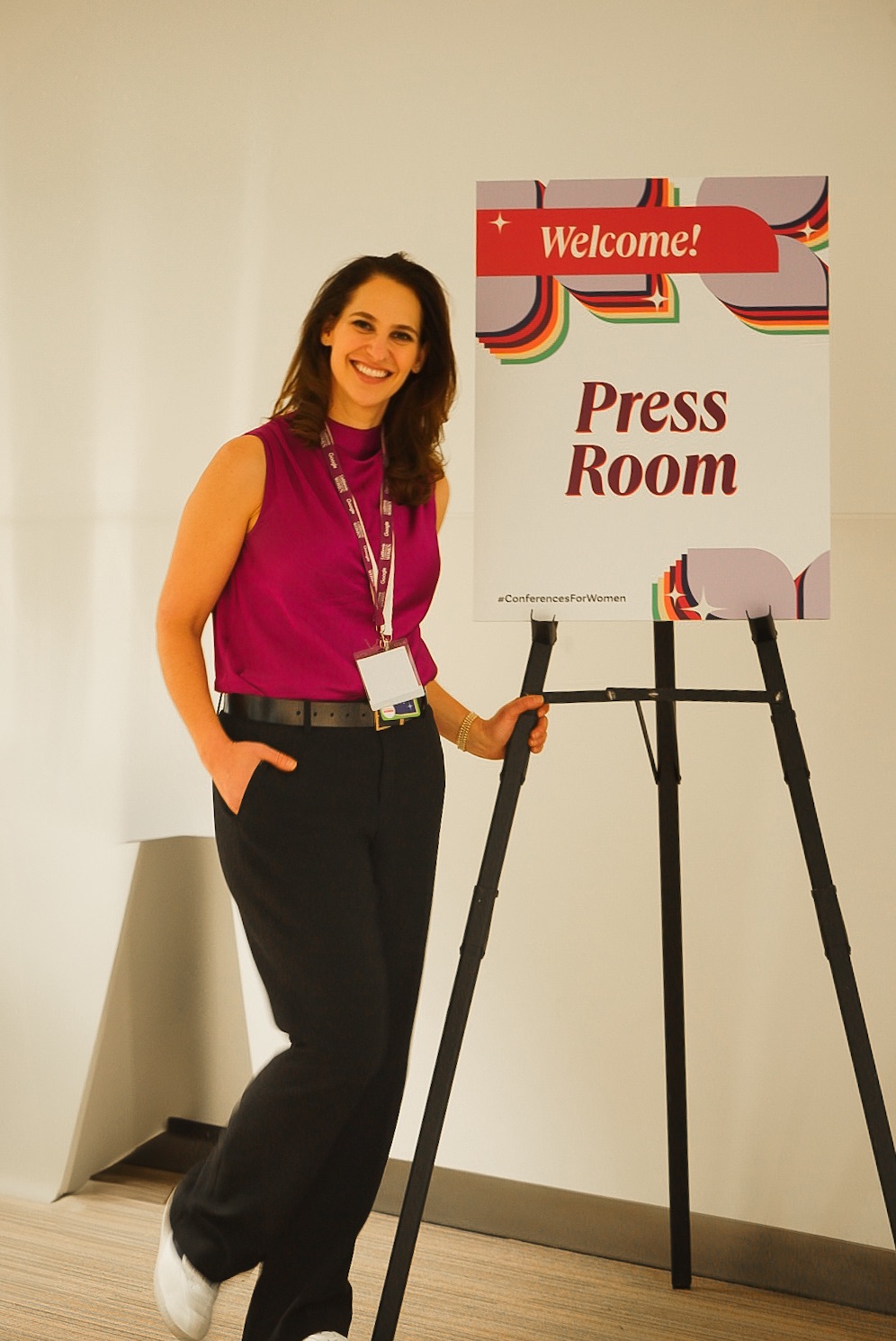 Woman standing next to a sign that says 'Press Room' and 'Welcome!', smiling, wearing a pink sleeveless top and black pants.
