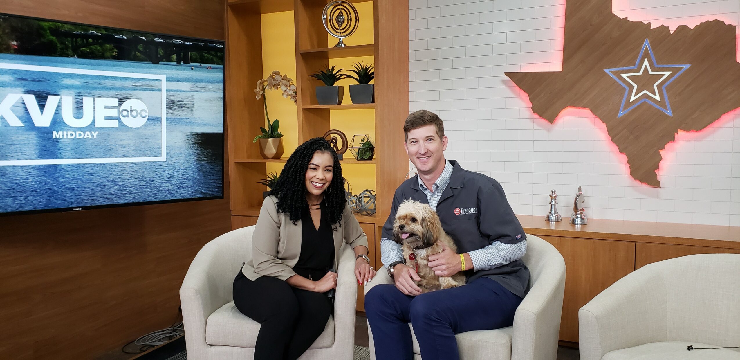 Two people sitting on a white couch, a woman and a man holding a small dog, in a TV studio setting.