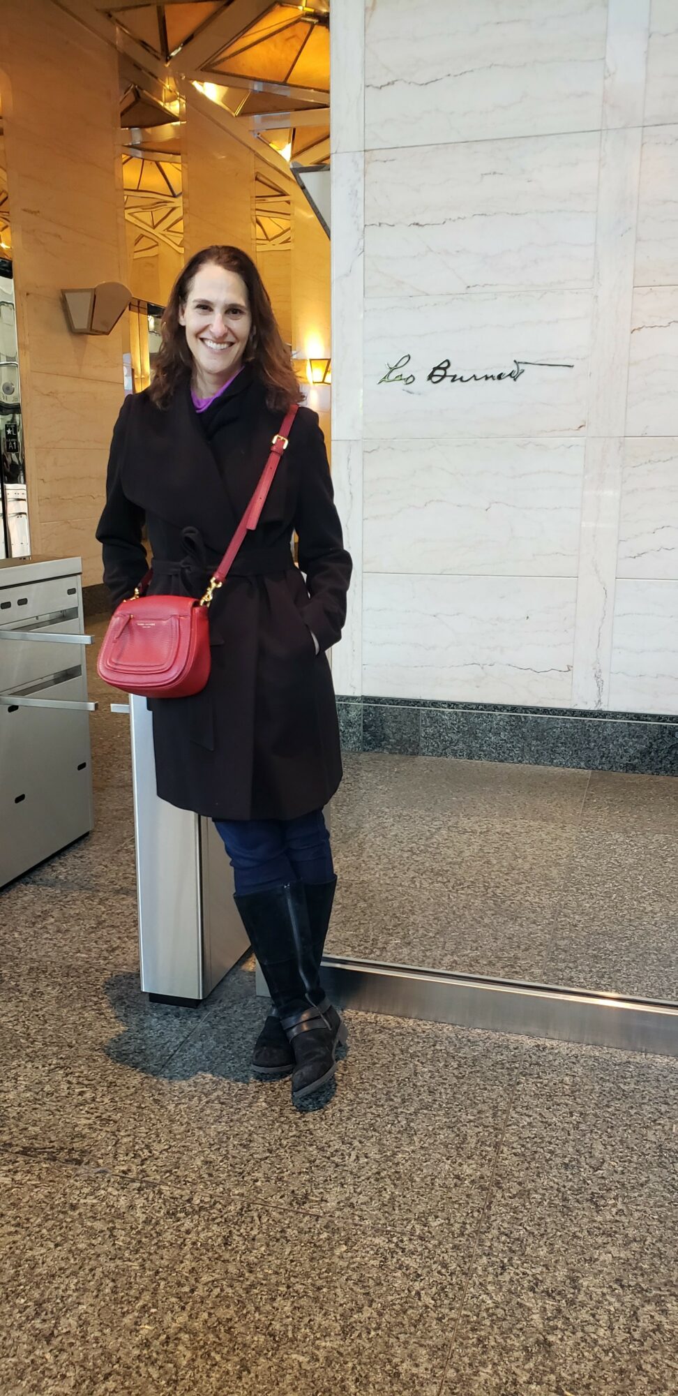 Woman standing indoors with a red purse, smiling, wearing a dark coat and jeans, in front of a white wall with writing.