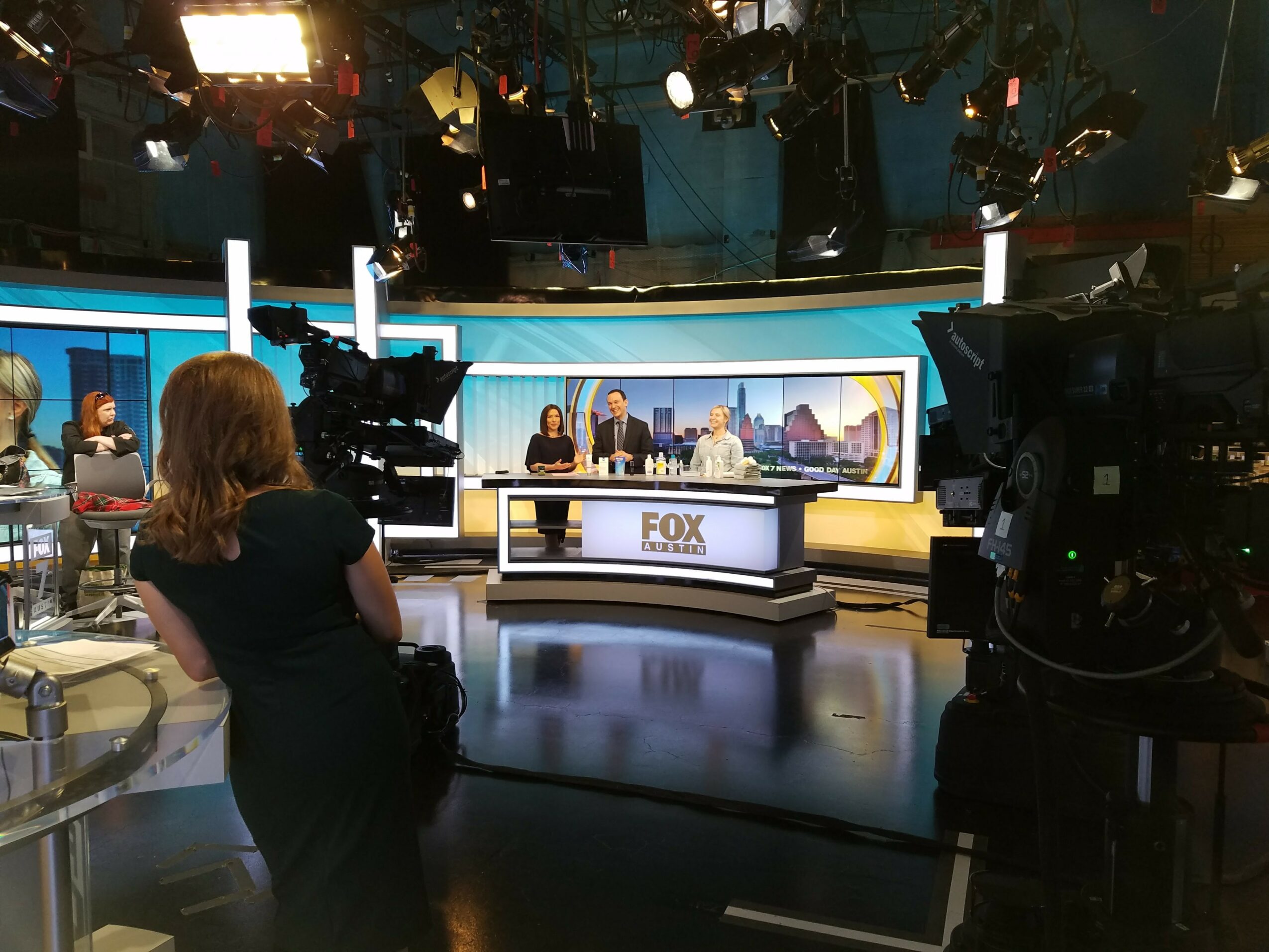 Television studio with news anchors at a desk, cameras, and crew members, bright studio lights, and a cityscape background on screen.