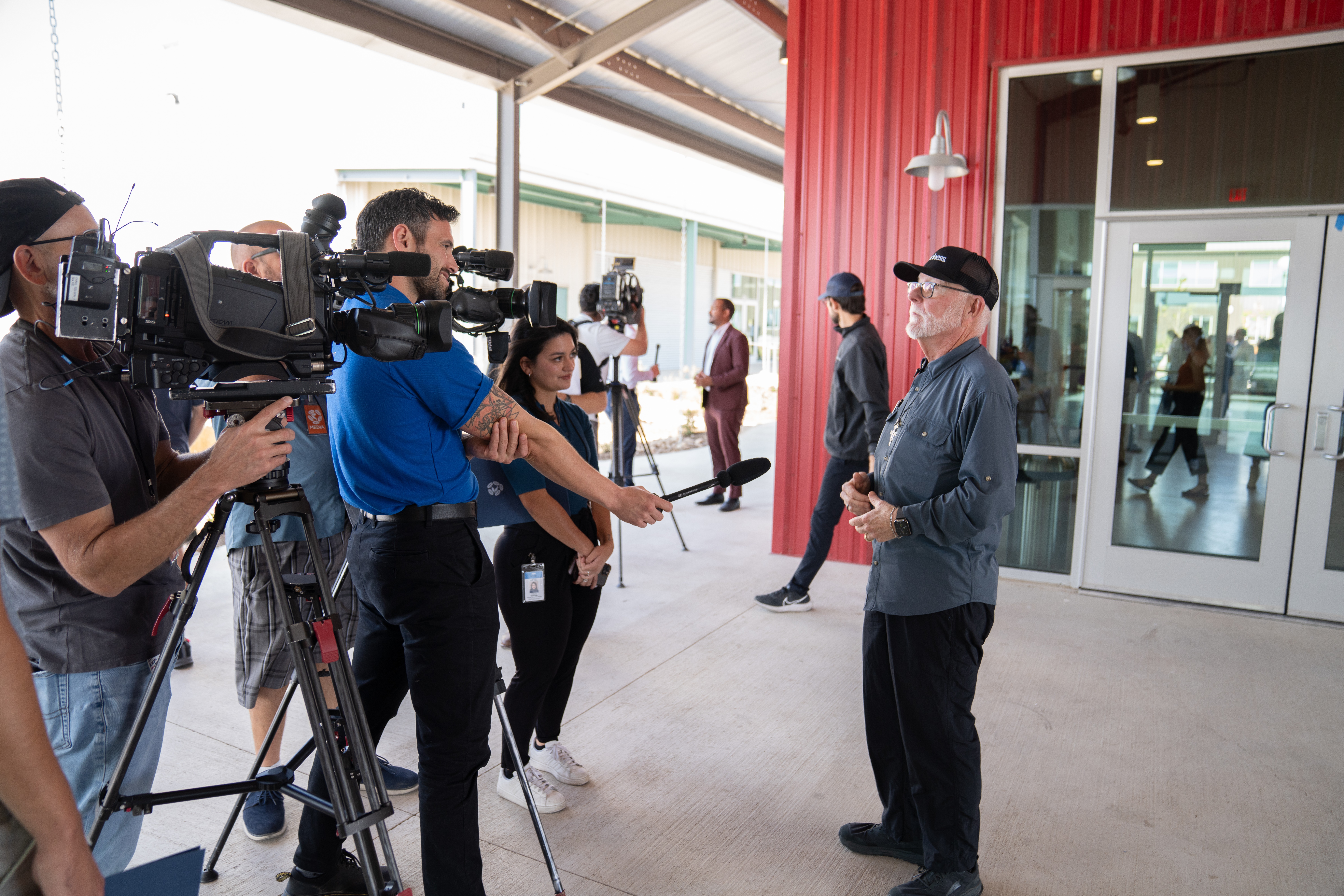 Group of people, including a person being interviewed, outside a building with glass doors and red wall, cameras filming.