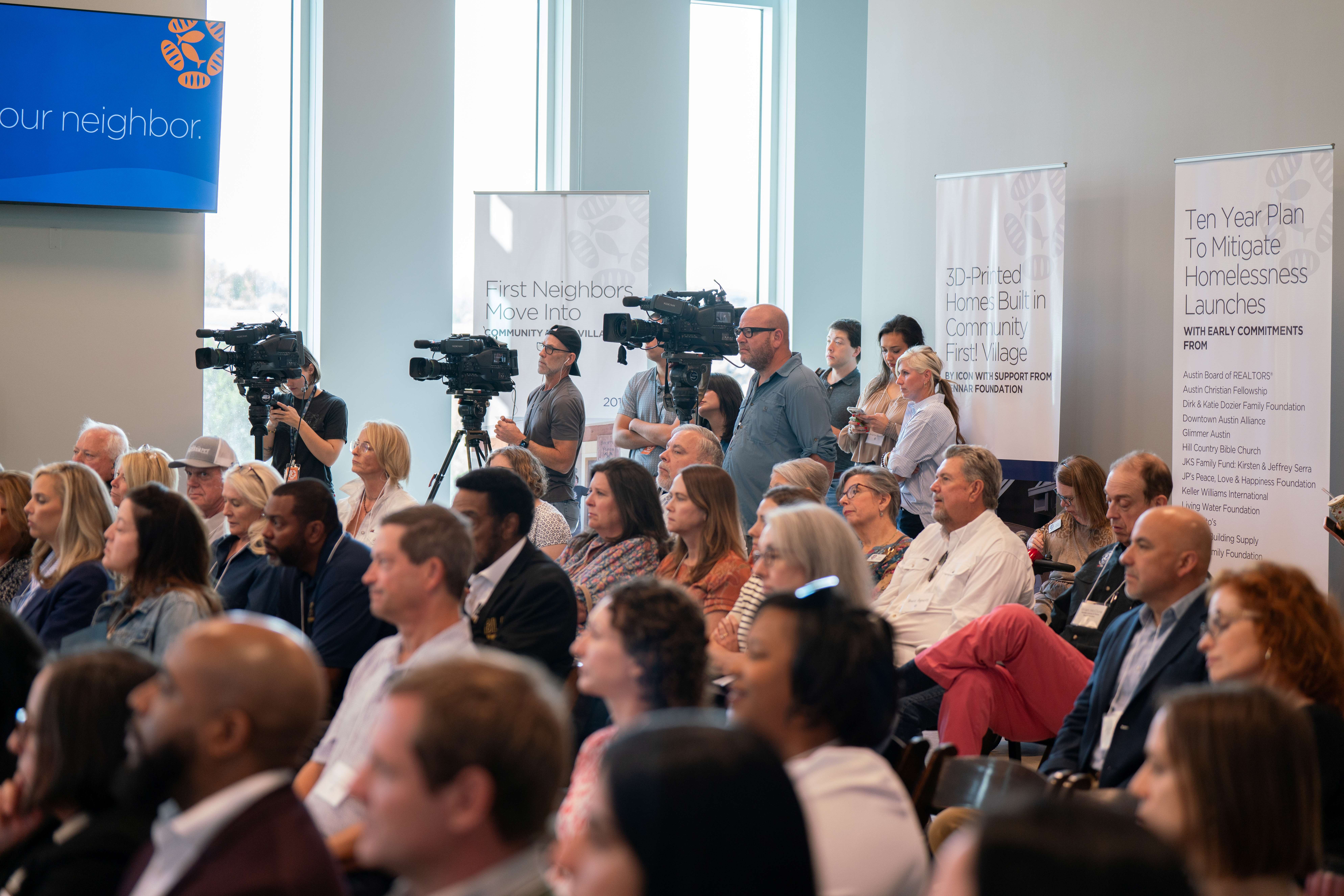 Audience seated in a conference room with cameras and banners, listening to a presentation or speech.