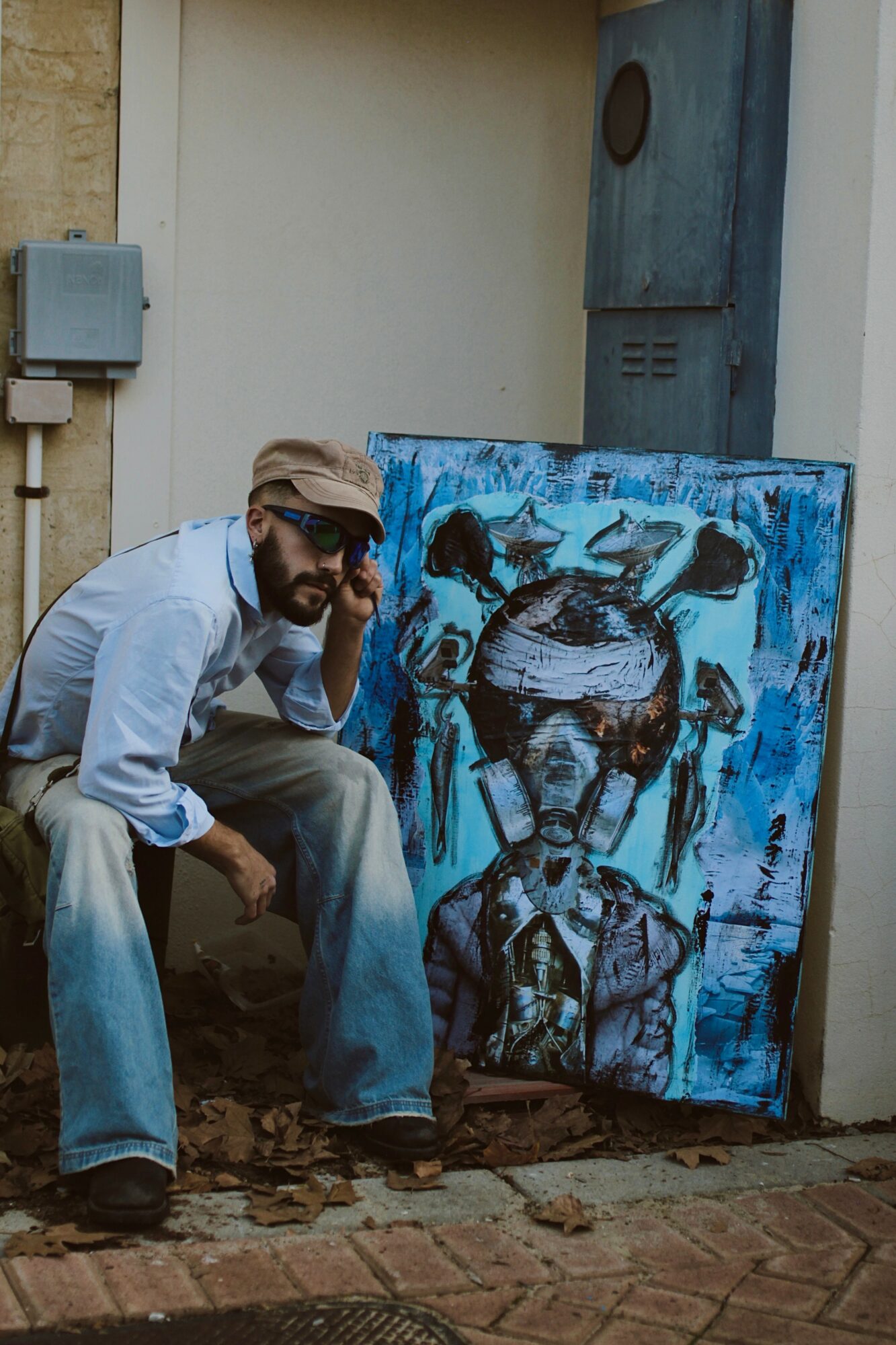 Man with sunglasses and beard crouching next to a colorful abstract painting outdoors, with a white wall and utility box behind.