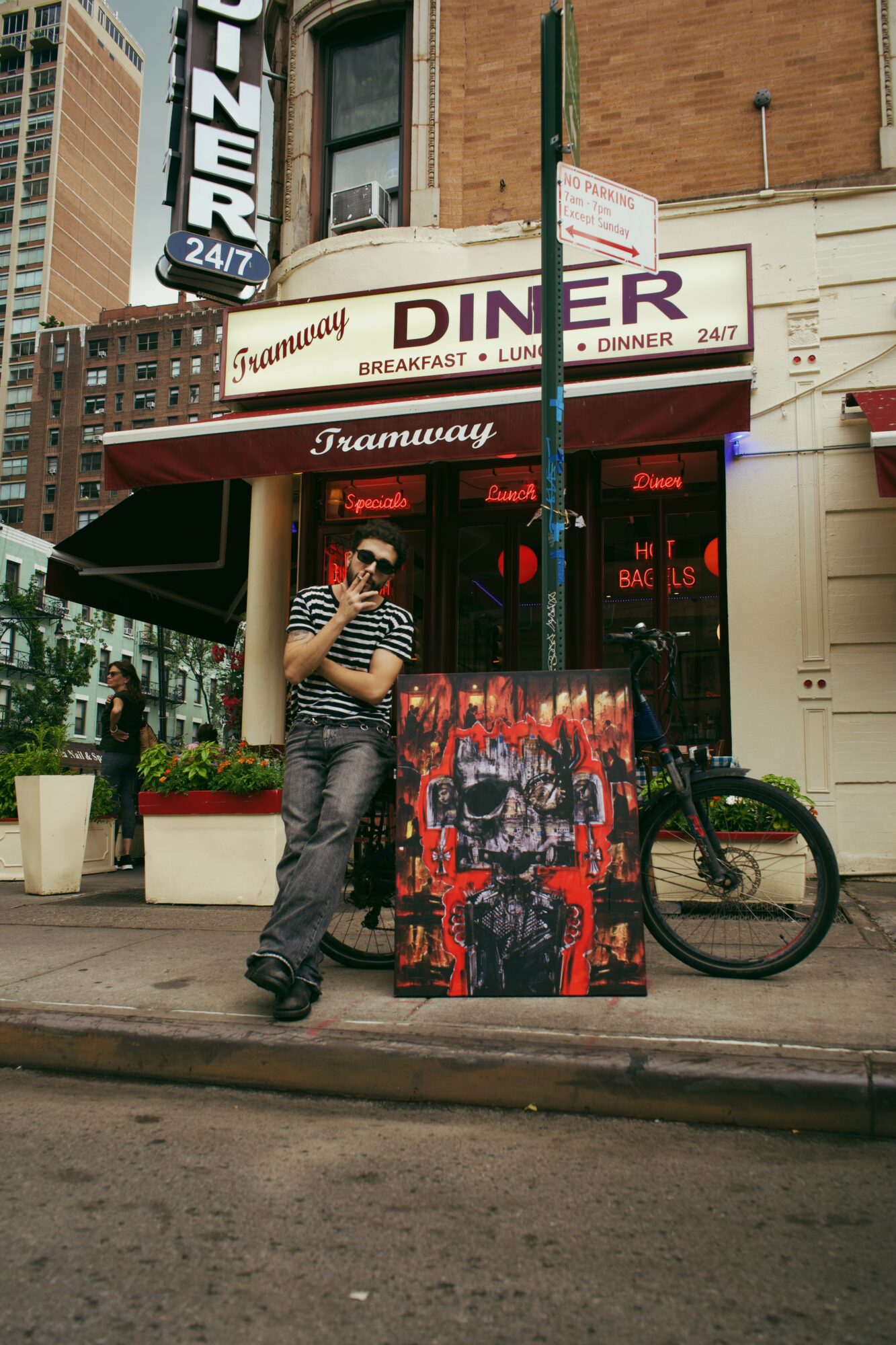 Person wearing striped shirt and sunglasses standing outside a diner with a red awning, bicycle nearby, urban street scene.
