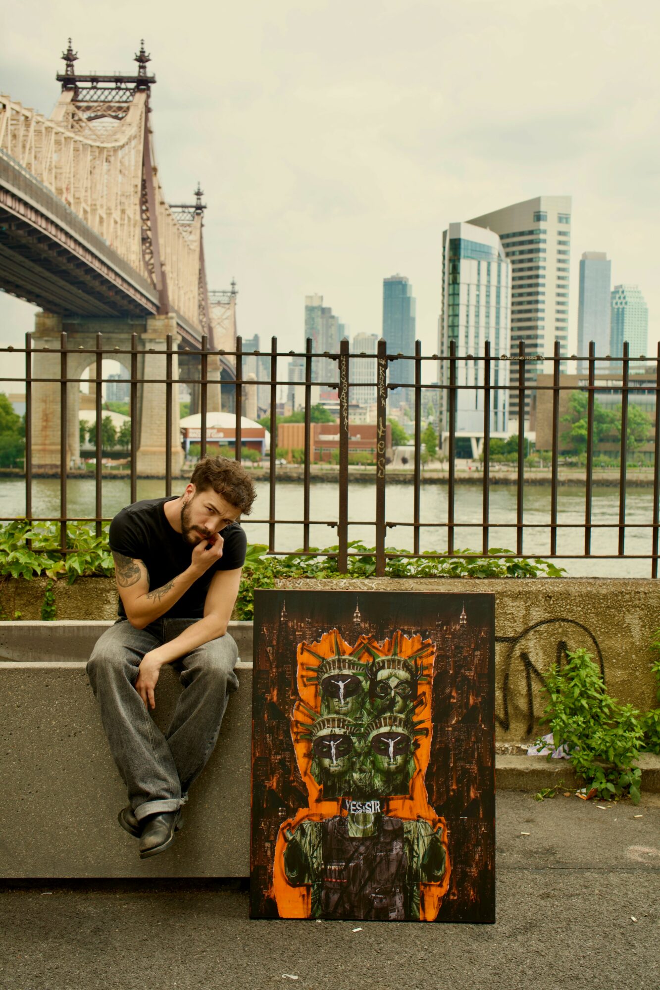 Person sitting on a ledge near a fence with a city skyline and bridge in background, next to a colorful painted artwork.