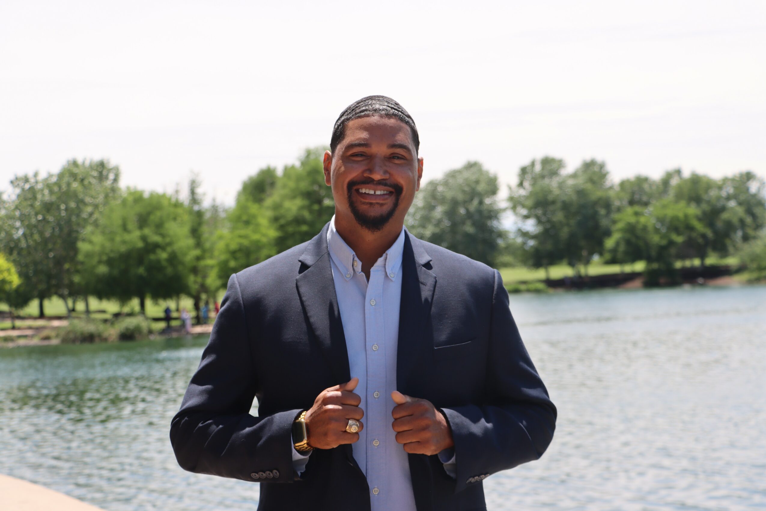 Man in a suit standing outdoors near a lake with trees in the background, smiling, holding his jacket lapels.