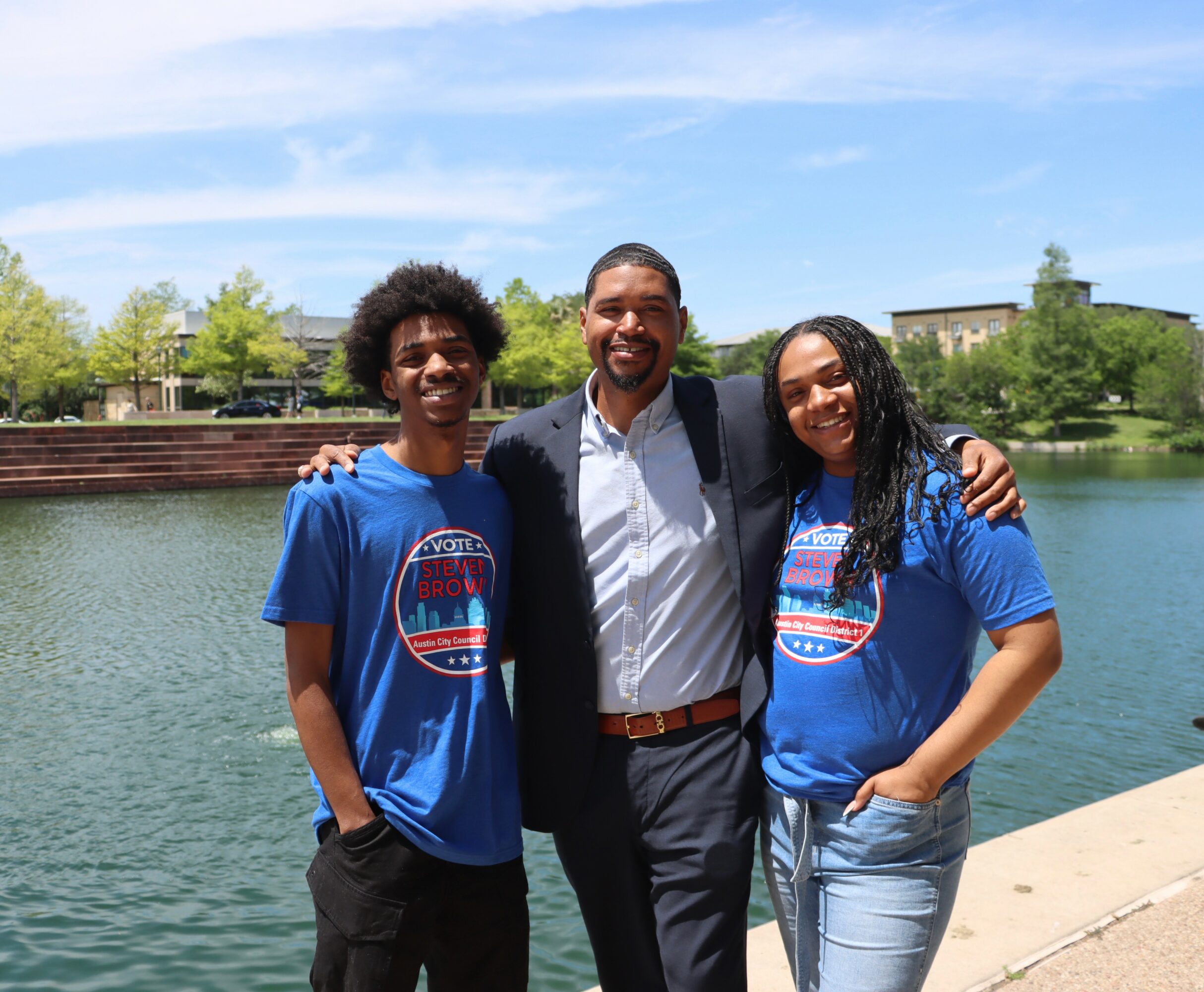 Three people standing outdoors near a body of water, smiling, with trees and buildings in the background.