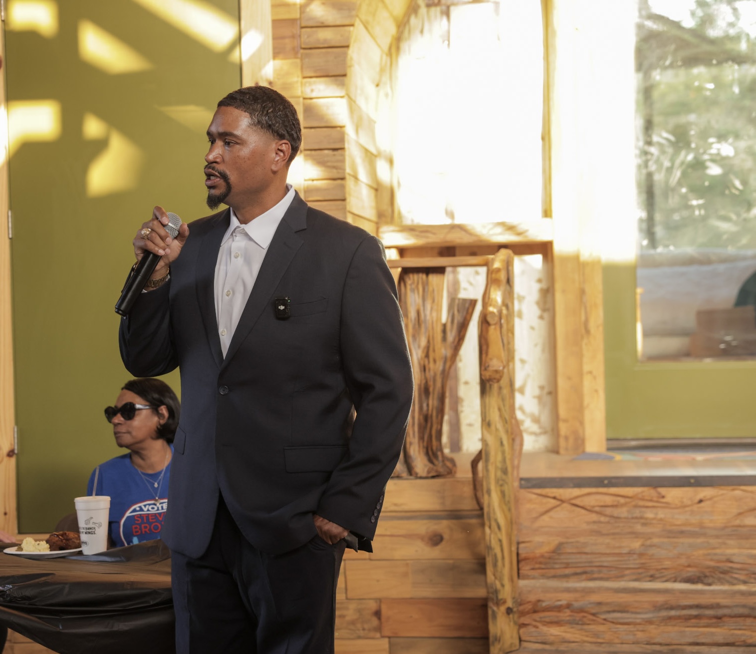 Man in suit holding microphone, standing indoors with wooden decor and a window, woman sitting at table in background.