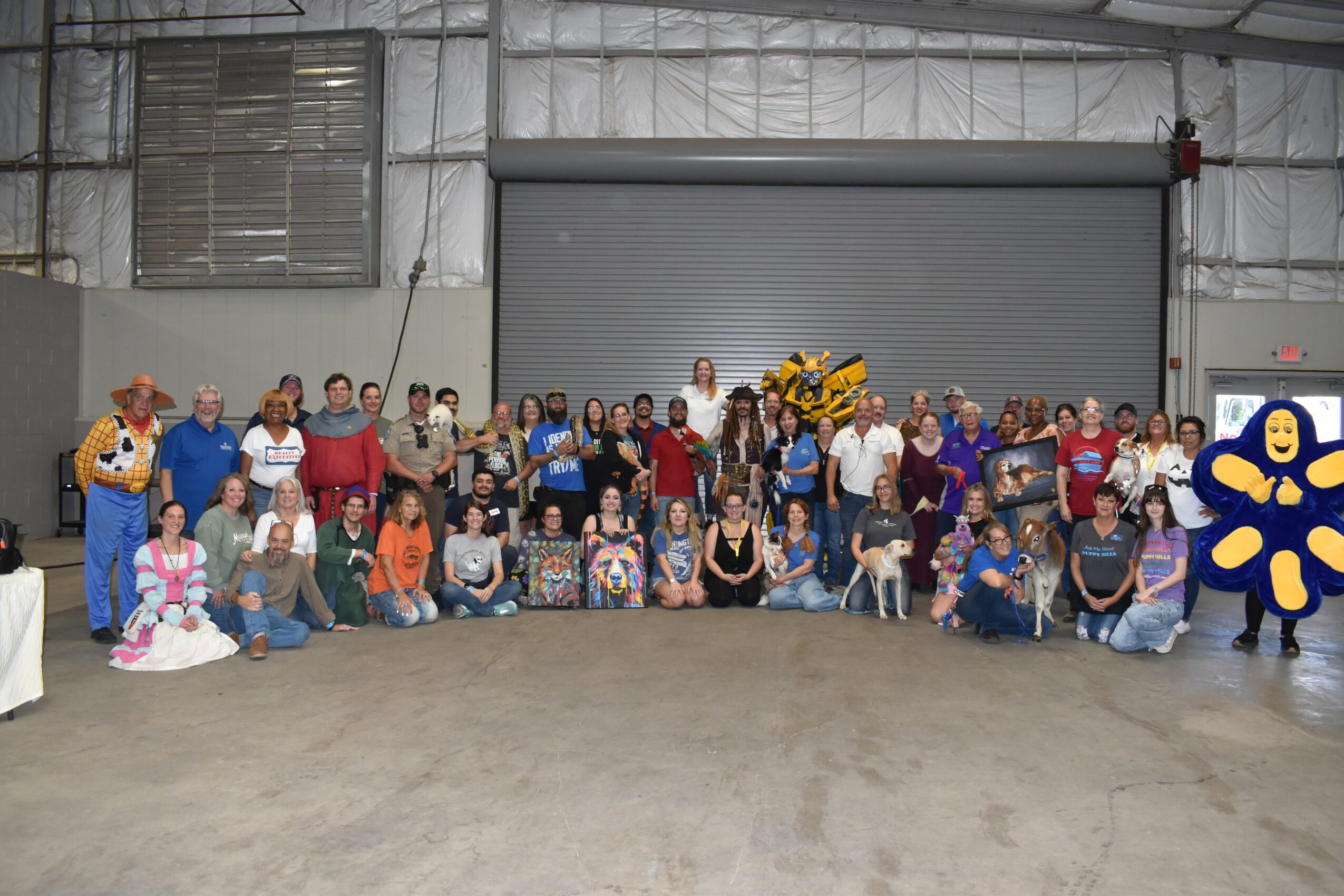 Large group of people gathered indoors in front of a closed garage door, with some decorations and a yellow flower arrangement.