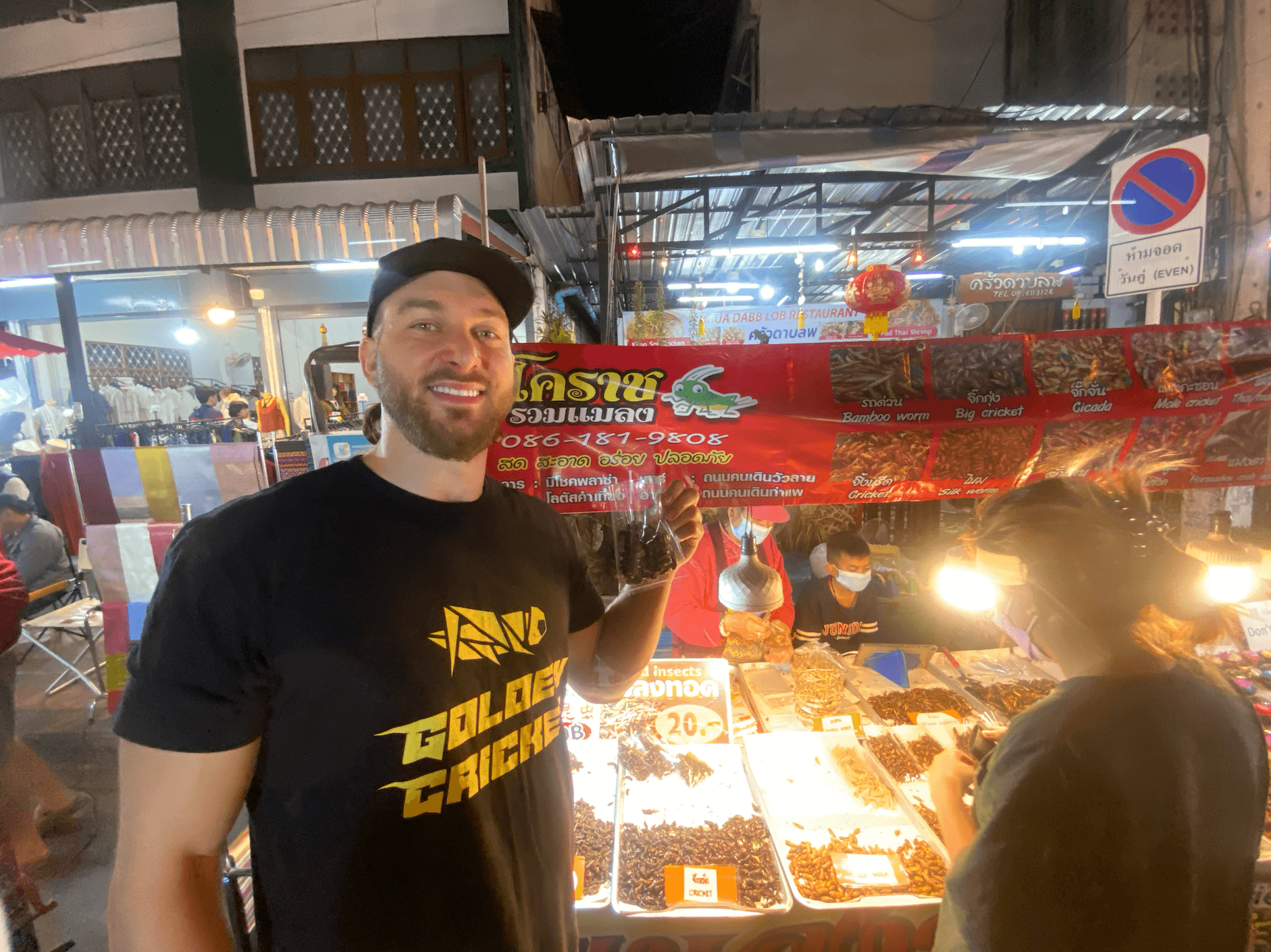 Man smiling holding a food item at a market stall with bright lights and people in background.