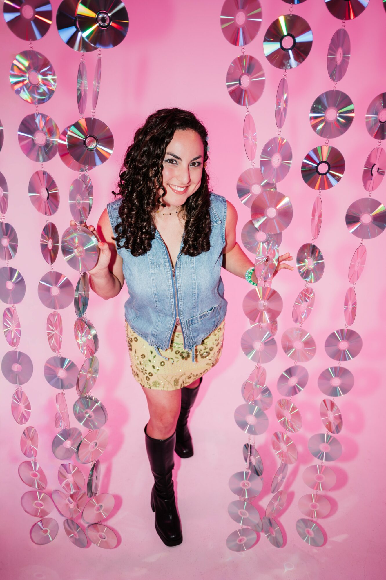 Young woman with curly hair stands among pink hanging decorations, wearing a denim vest, yellow skirt, and black boots.