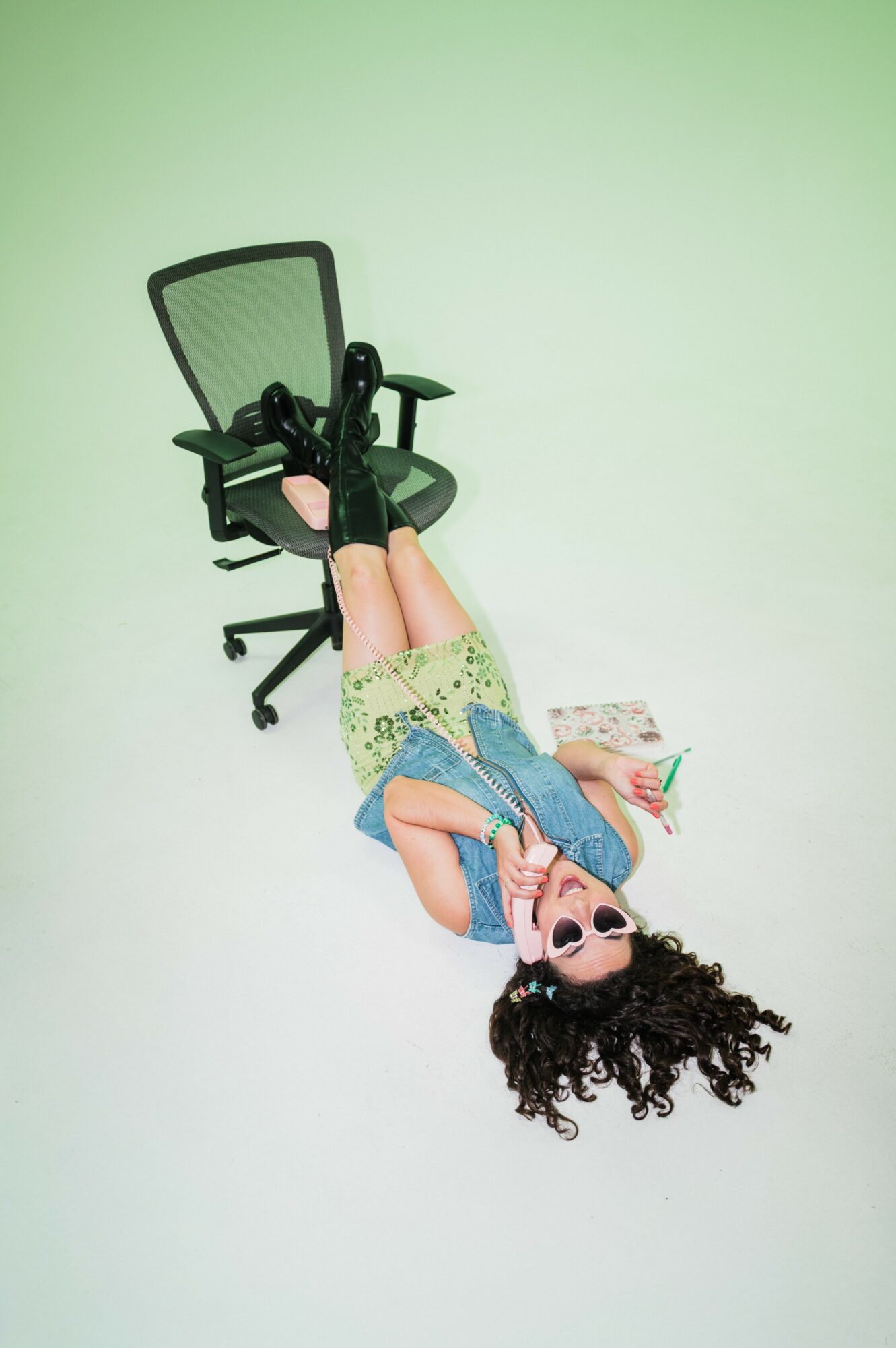Child lying on the floor with a chair behind, wearing sunglasses, a blue shirt, and patterned shorts, smiling.