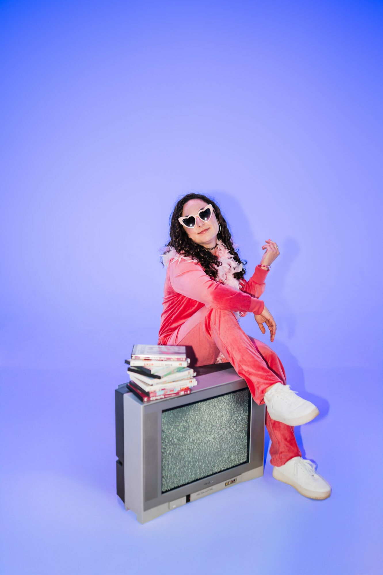 Person sitting on a vintage TV with books, wearing sunglasses and a pink outfit, against a blue background.