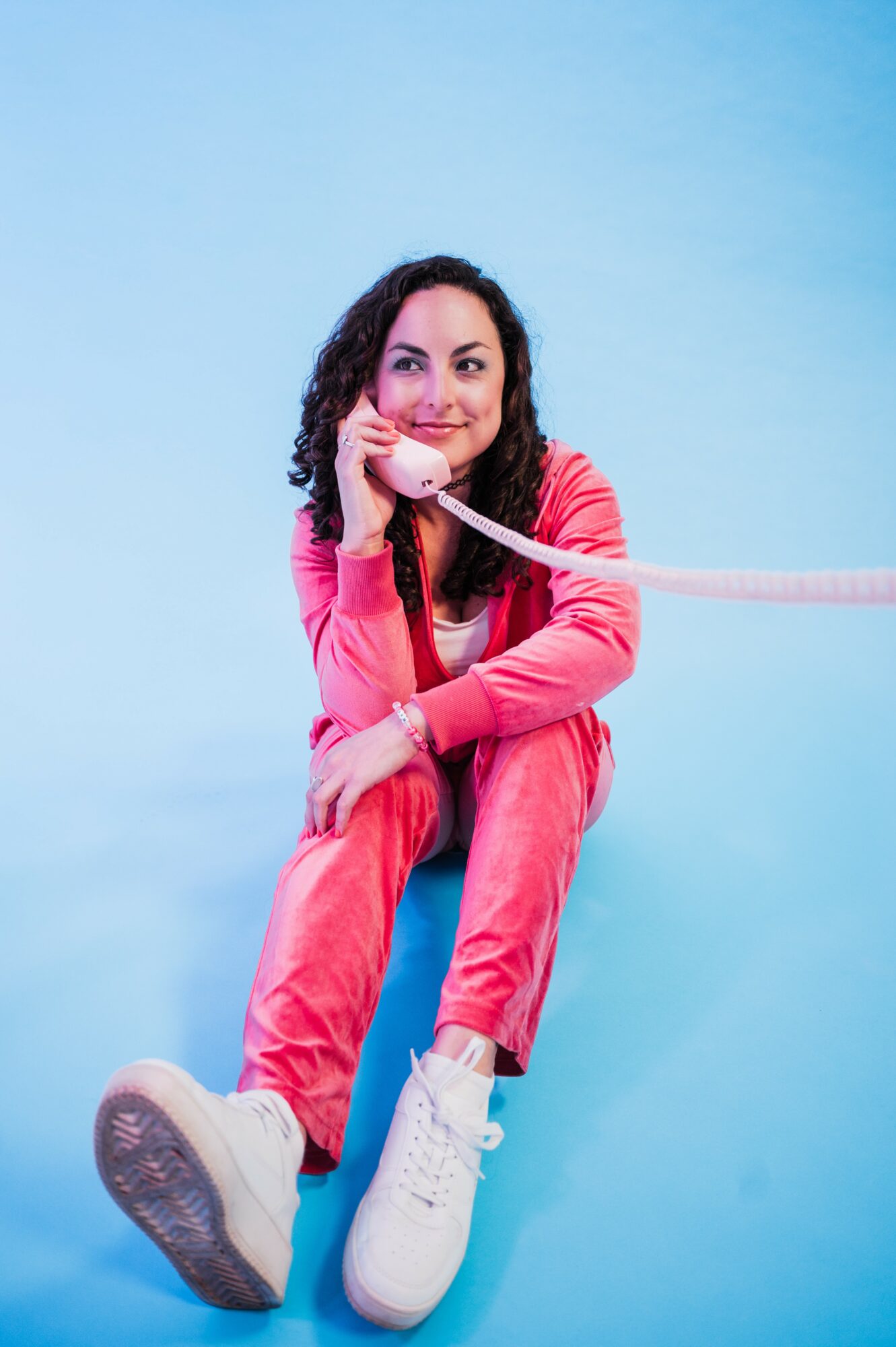 Young woman with curly hair sitting on a blue chair, holding a pink telephone receiver, smiling, against a light blue background.