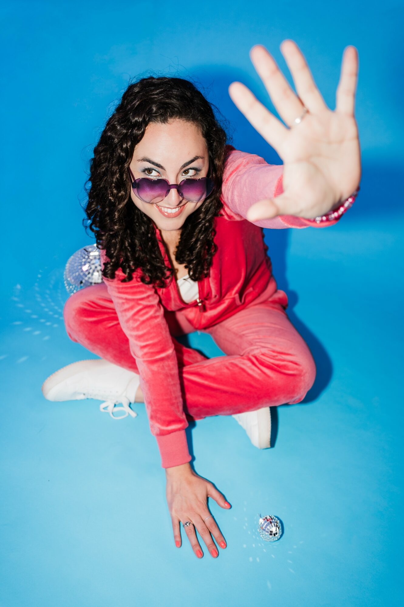 Woman with curly hair, glasses, and pink outfit reaching out with hand, smiling, against blue background.