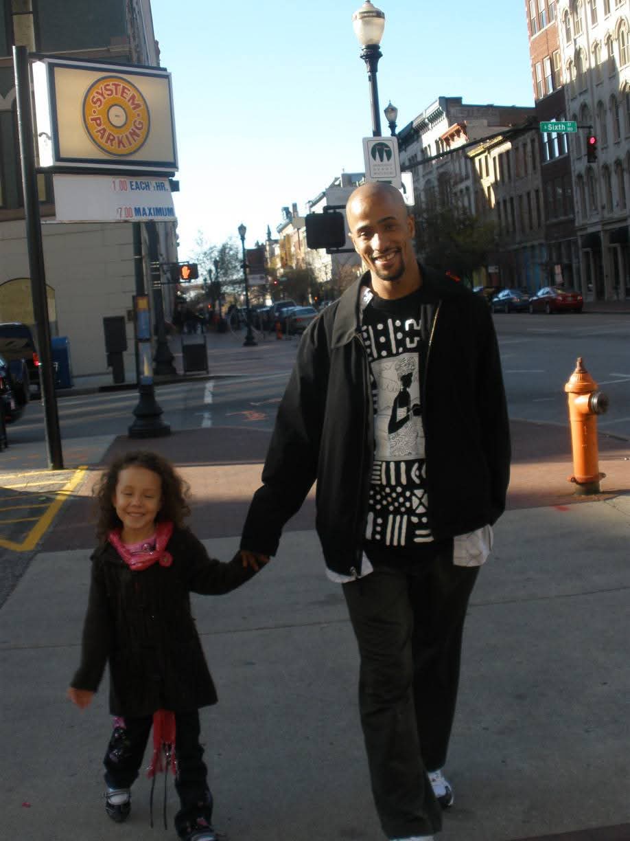 A man and a young girl holding hands walking on a city street, smiling, with buildings and a traffic light in the background.