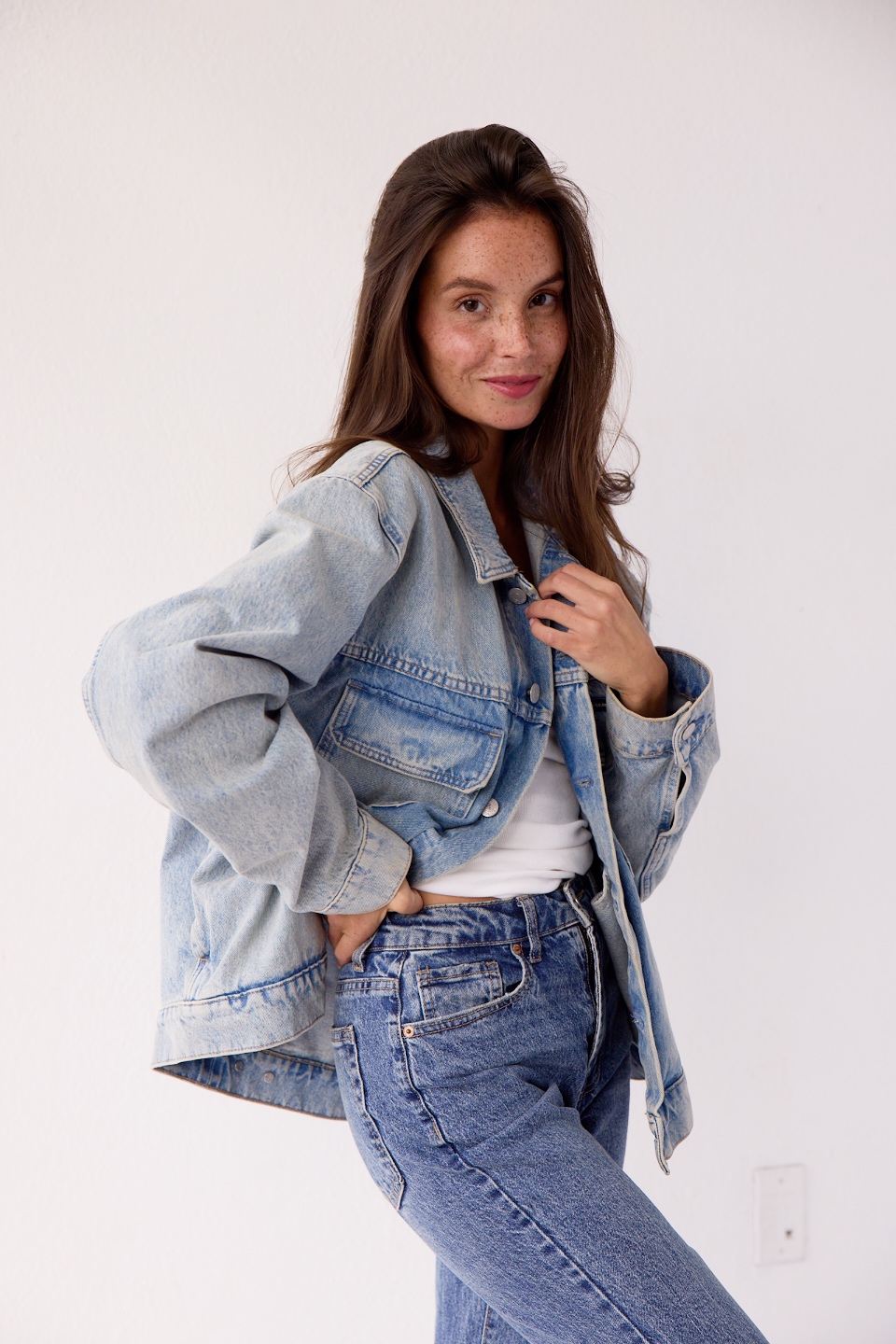 Young woman with long brown hair wearing a denim jacket and jeans, smiling, standing against a plain white background.