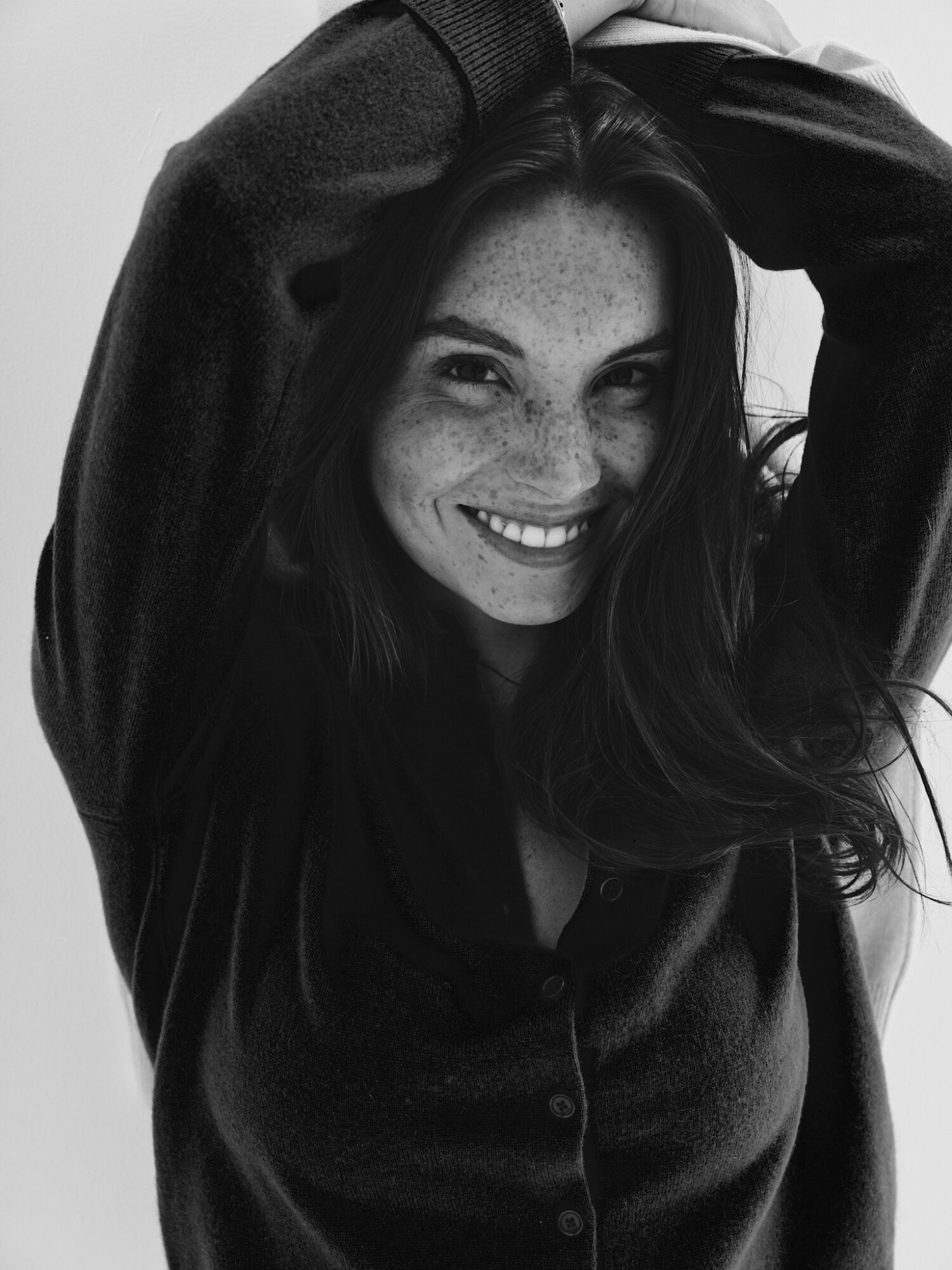 Smiling woman with long hair, hands behind head, wearing dark clothing, in black and white.