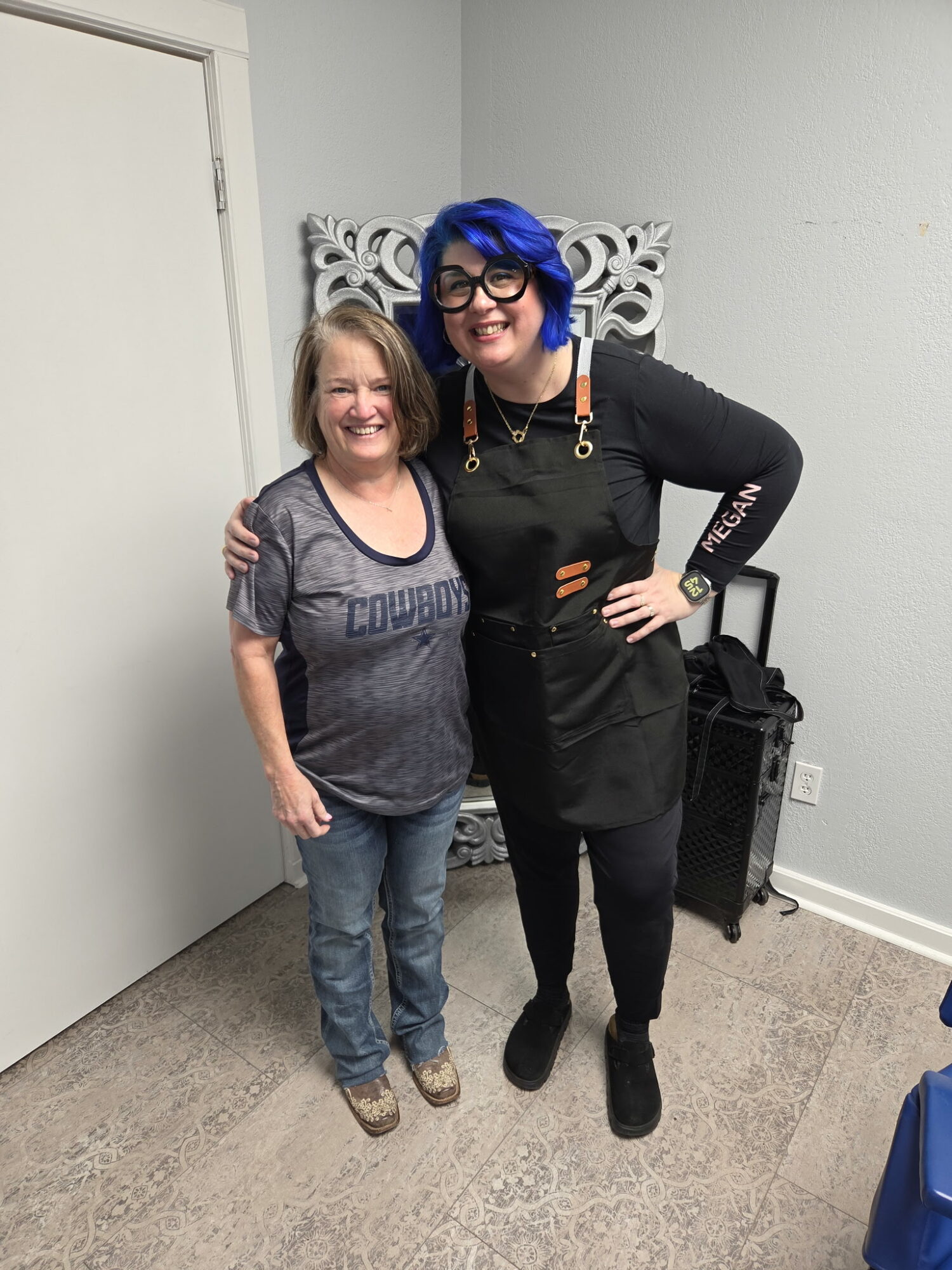 Two women standing together, smiling, one with blue hair and glasses, the other with blonde hair, in a room with gray walls.