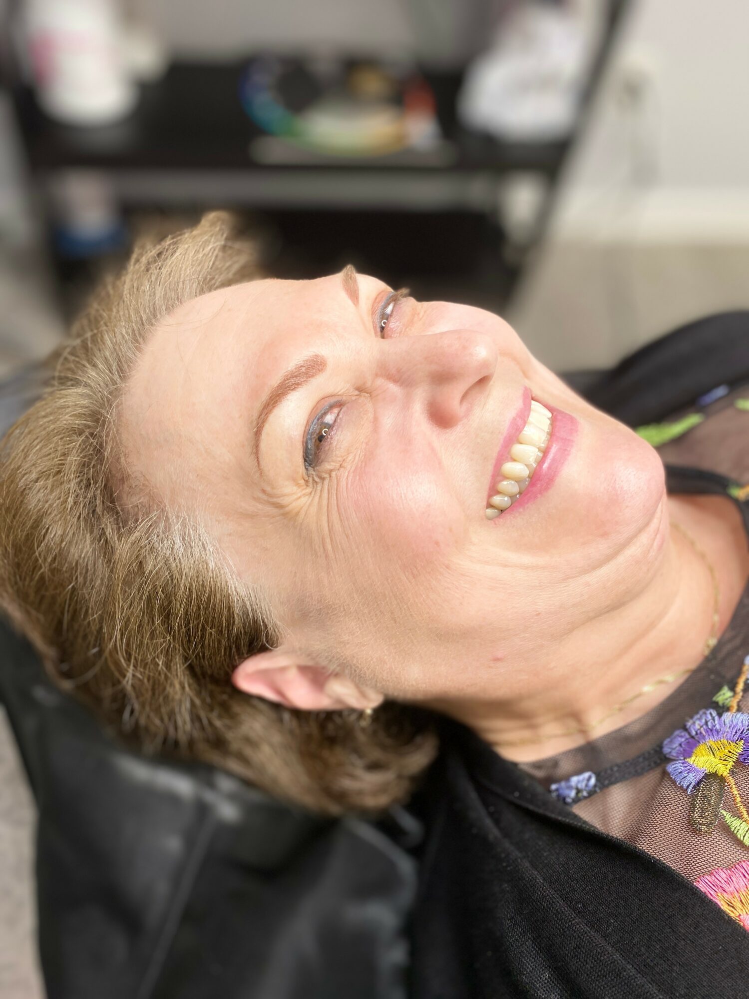 Woman lying down, smiling, with a background of a shelf and objects, wearing a black top and colorful necklace.