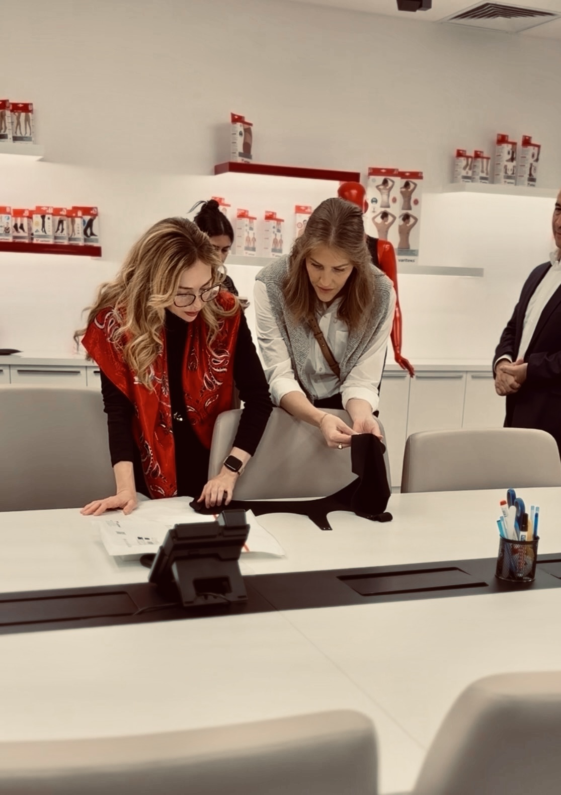 Two women leaning over a table, examining documents, in a room with shelves of red and white items, and a person standing nearby.