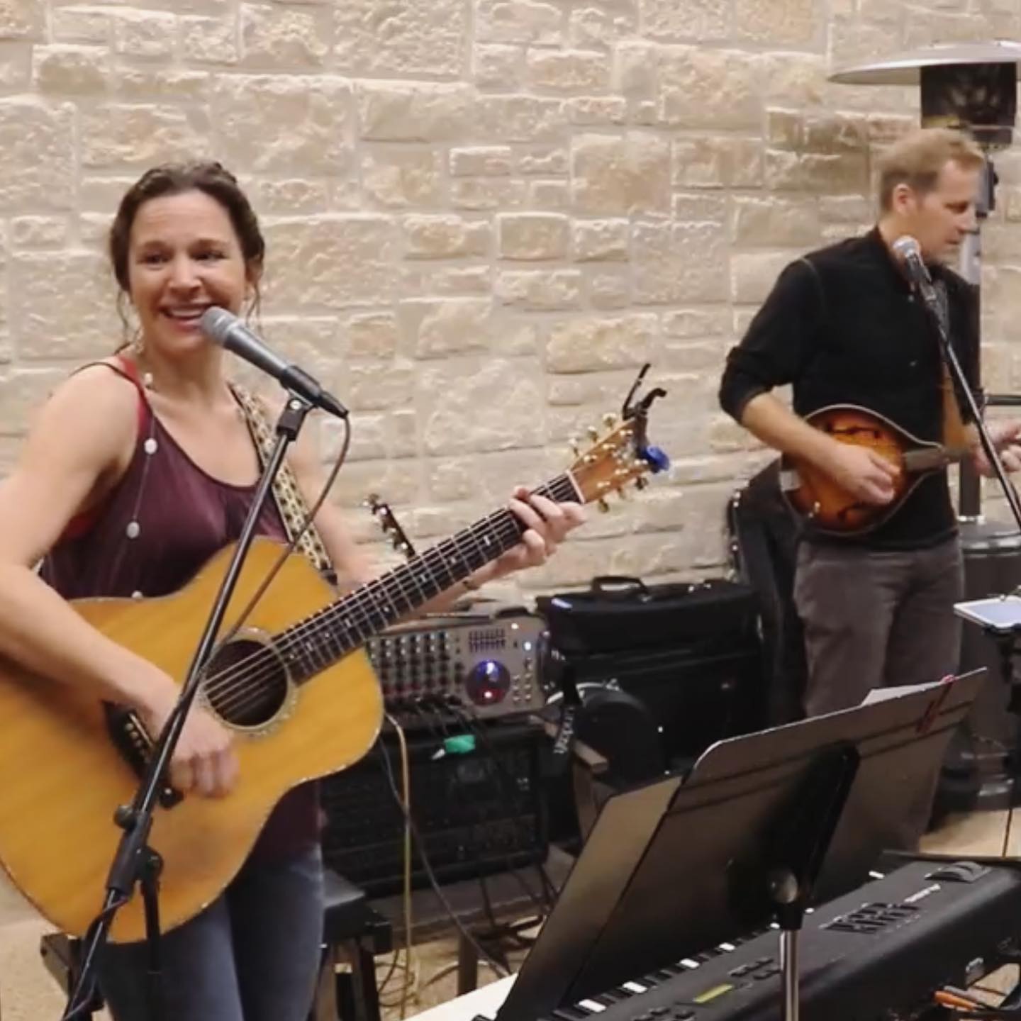 Two women playing guitars and singing, one smiling at microphone, in a room with a brick wall background.