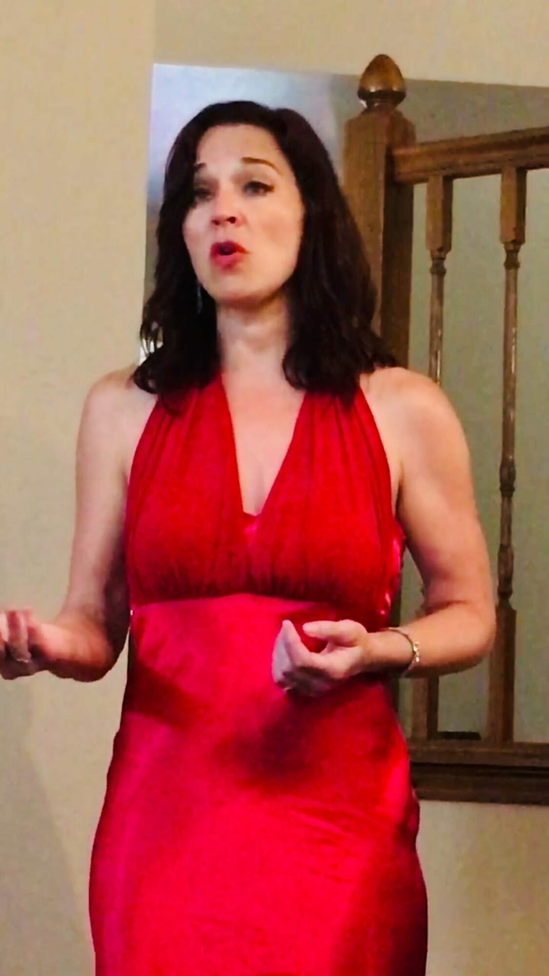 Woman in red dress standing indoors, gesturing with her hands, with a staircase in the background.