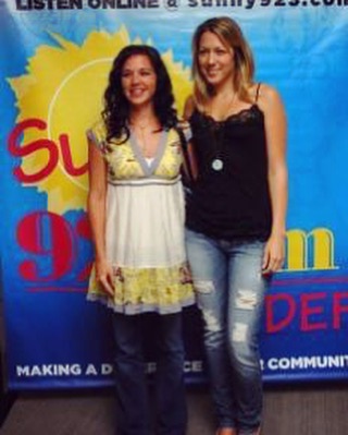 Two women standing in front of a colorful backdrop, smiling, with one wearing a white and yellow dress and the other in a black top and ripped jeans.