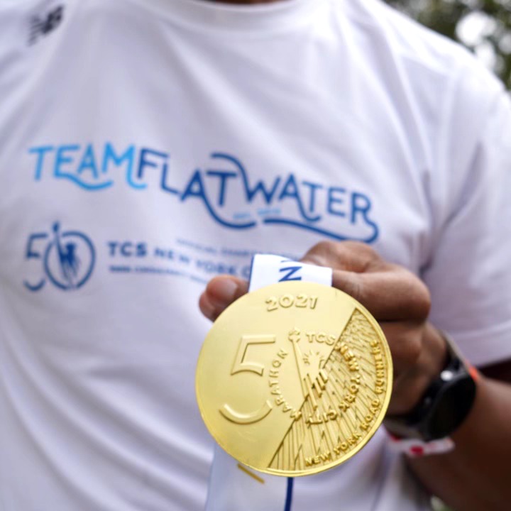 Person holding a large gold 5 euro coin in front of a white T-shirt with blue text. Background is outdoors.