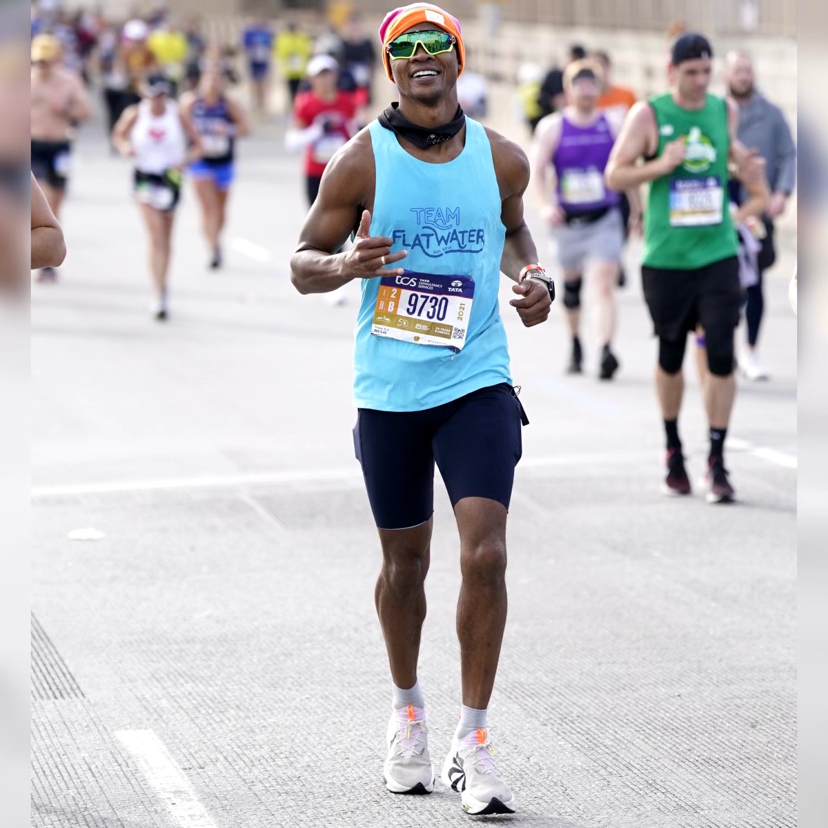 Runner in blue tank top and black shorts smiling during a race, with other runners in the background.
