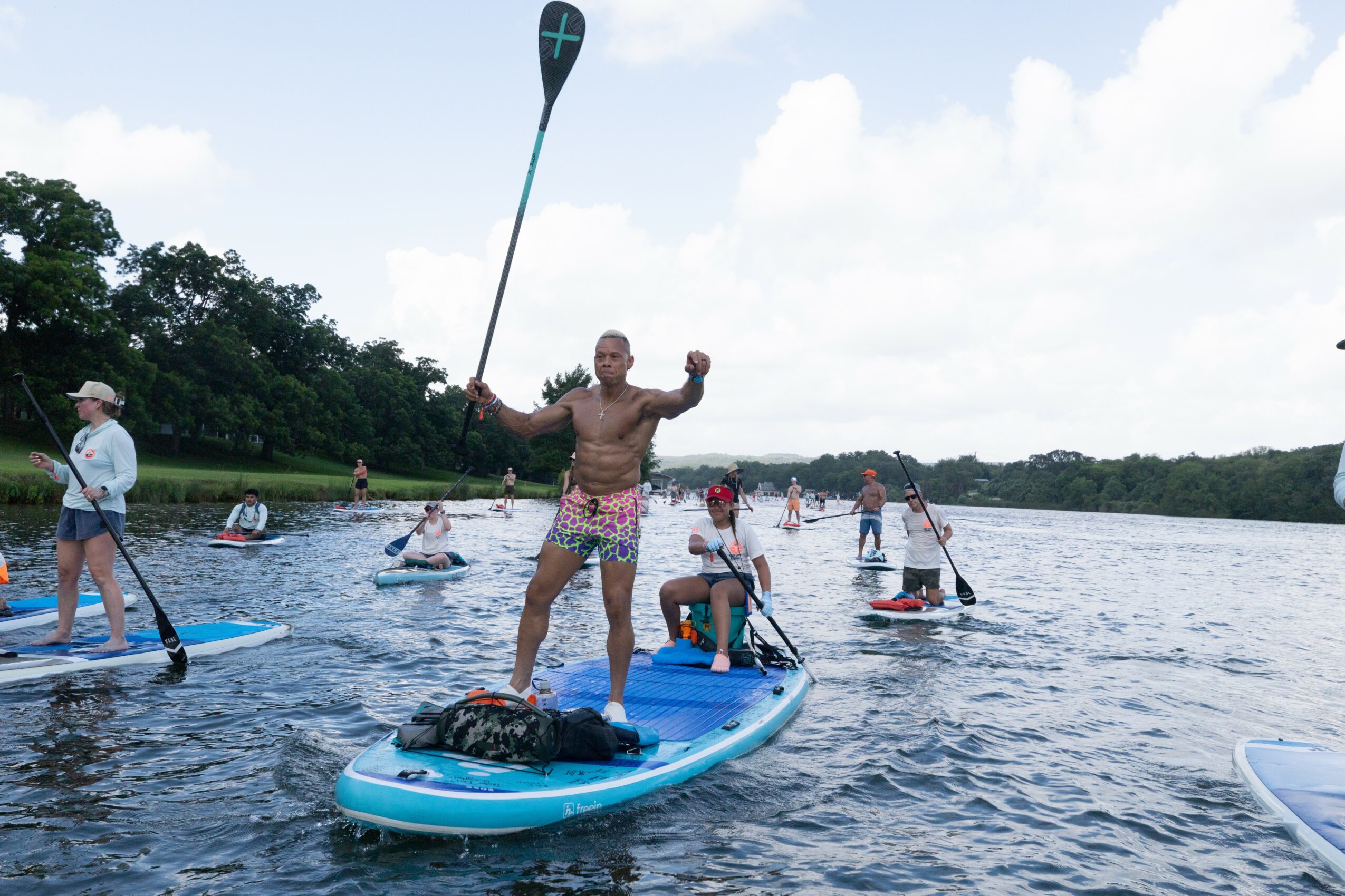 Person standing on paddleboard holding paddle, surrounded by others on paddleboards and kayaks on water, trees in background.