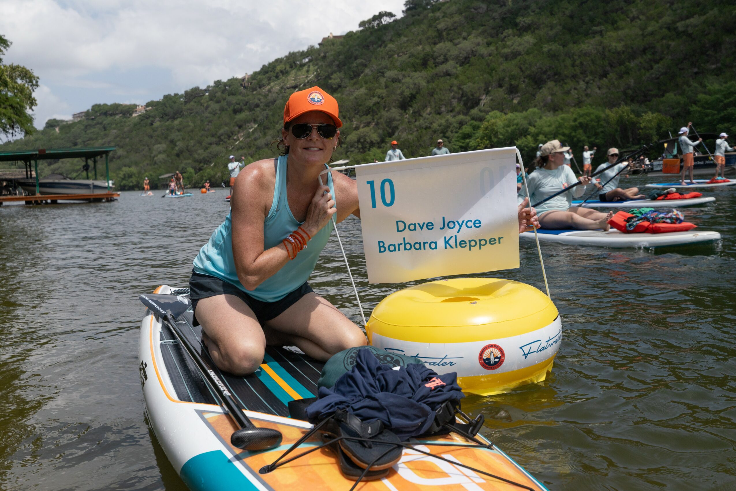 Woman in sunglasses and orange cap on paddleboard holding a sign with names, surrounded by water and boats.