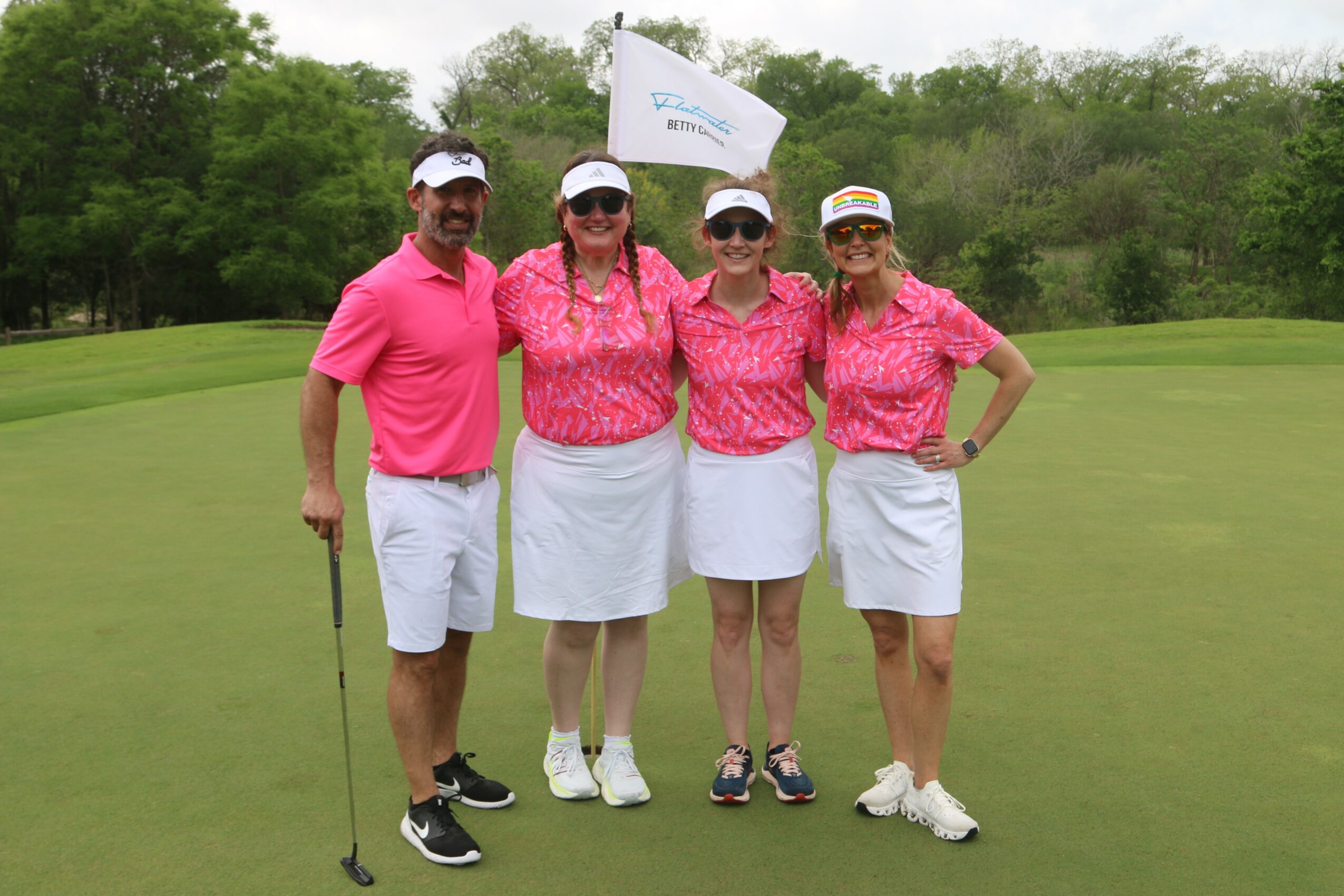 Four women in pink shirts and white skirts standing on a golf course, with a flag and trees in background.