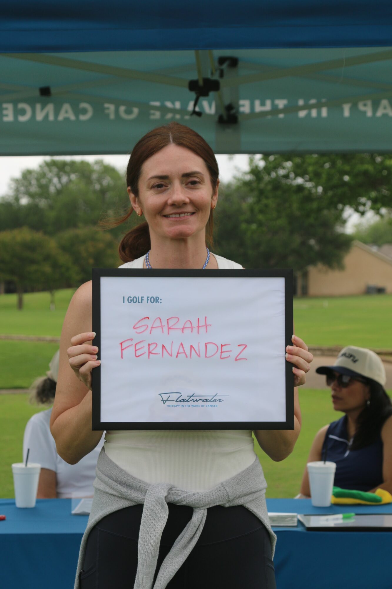 Woman holding a framed certificate outdoors, smiling, with people and a green field in the background.