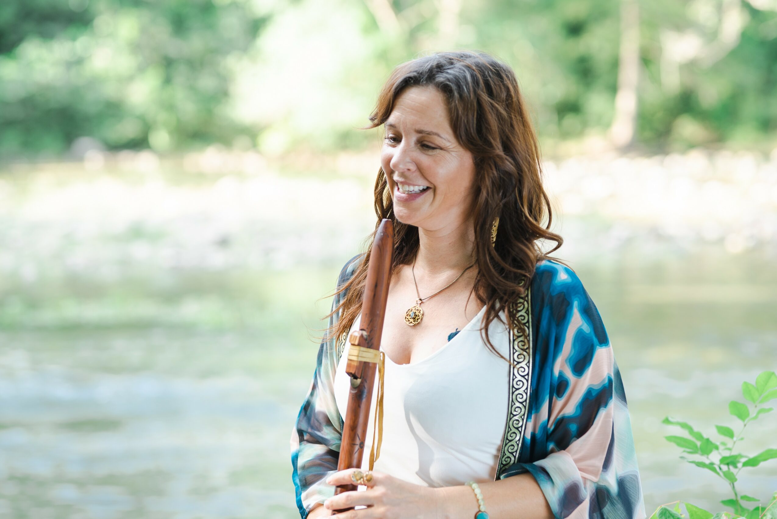Woman smiling outdoors near a river, wearing a white top and a blue patterned shawl, with trees in the background.