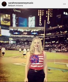 Young woman with blonde hair smiling at a baseball stadium, wearing a striped top with a floral pattern, standing on the field.
