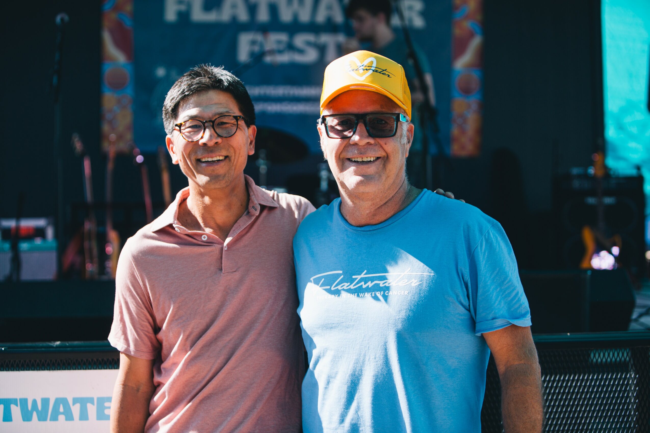Two men smiling, standing outdoors with a stage and banner in the background.