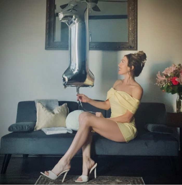 Woman in yellow dress sitting on sofa holding a balloon shaped as the number one, with flowers nearby.