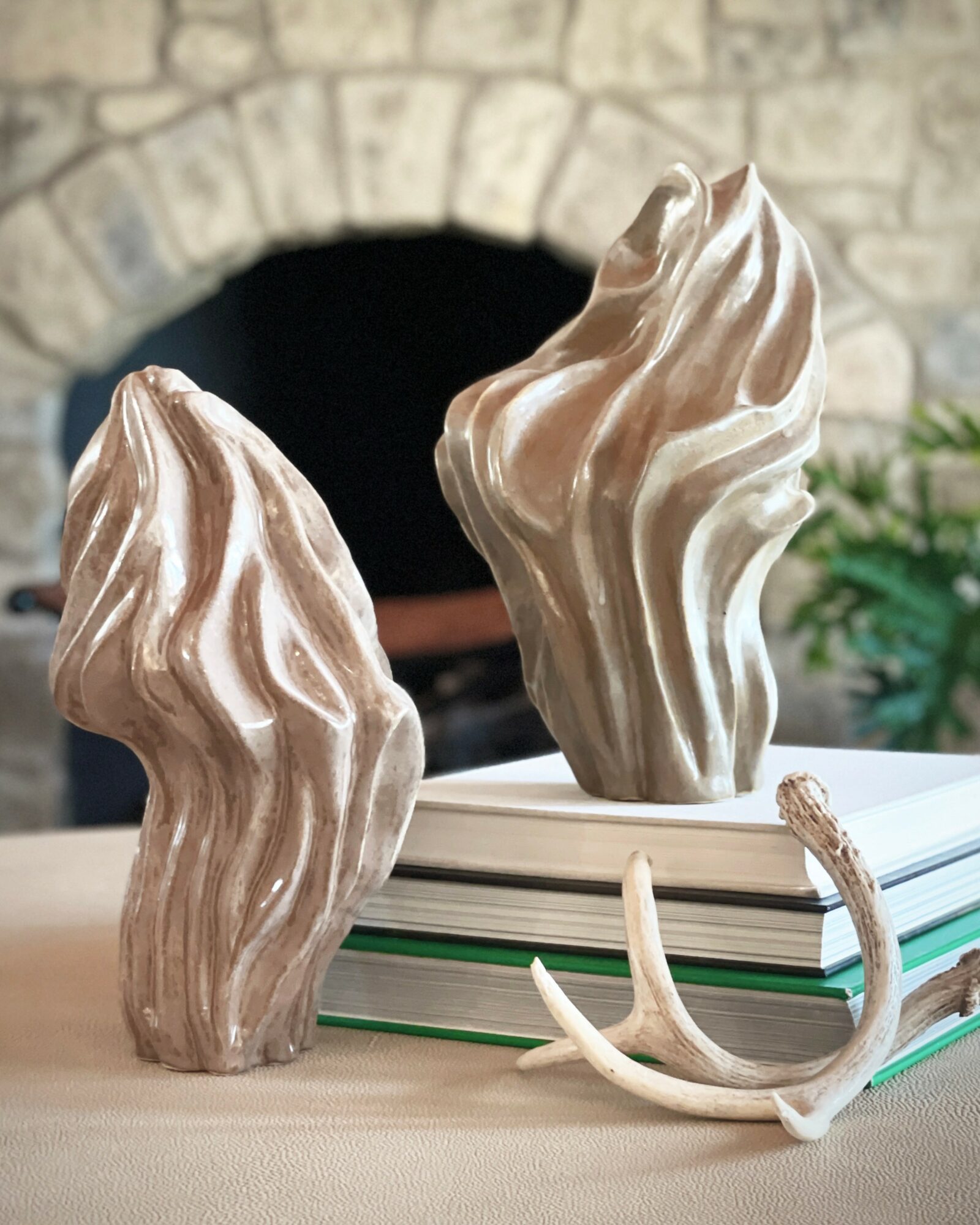 Two large, sculptural objects resembling twisted, textured rocks or wood, placed on a stack of books with a stone fireplace in the background.