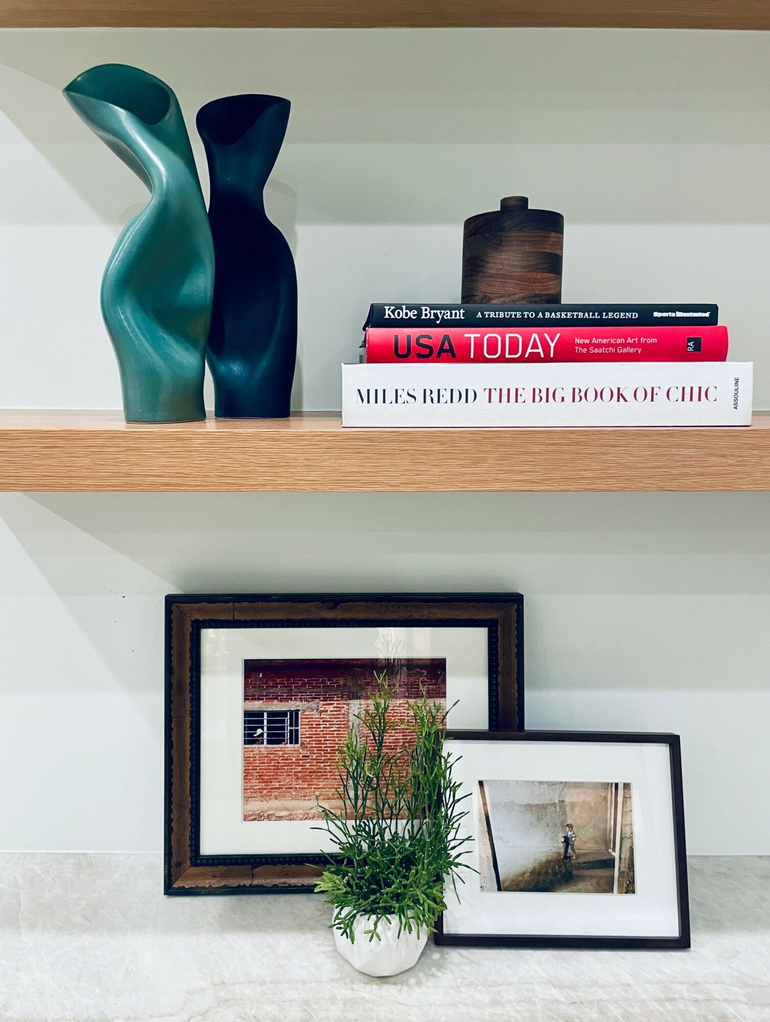 Shelf with two vases, books, framed photos, and a potted plant, arranged on a white wall, viewed from the front.