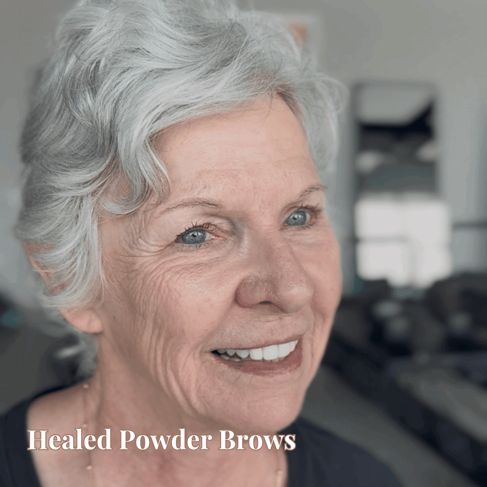 Close-up of smiling elderly woman with short, curly gray hair and healed powder brows, in a bright room.