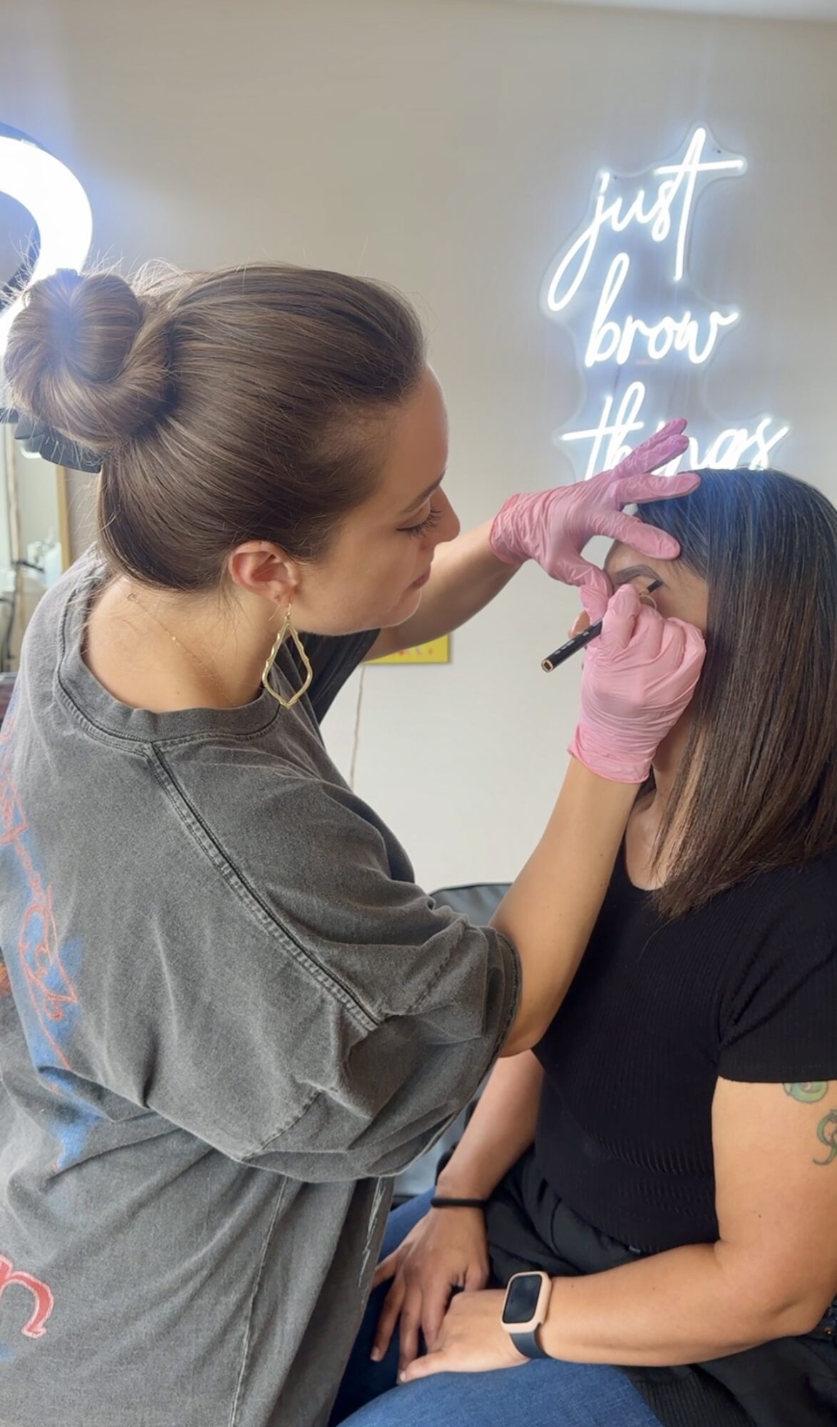 A woman with a bun and pink gloves performs a cosmetic procedure on another woman with dark hair, in a salon setting.