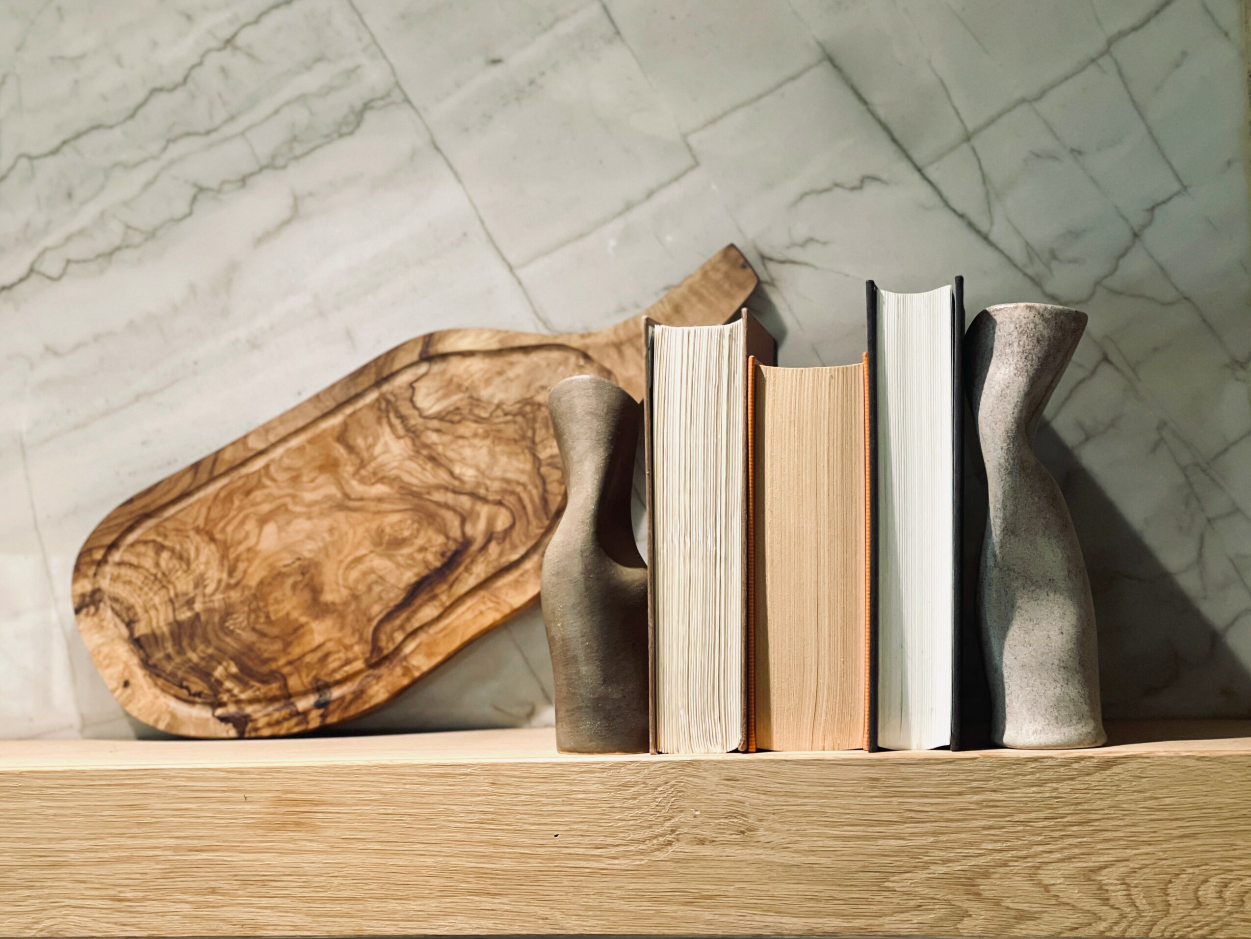 Books and vases on a wooden shelf against a marble wall, with a wooden tray behind them.