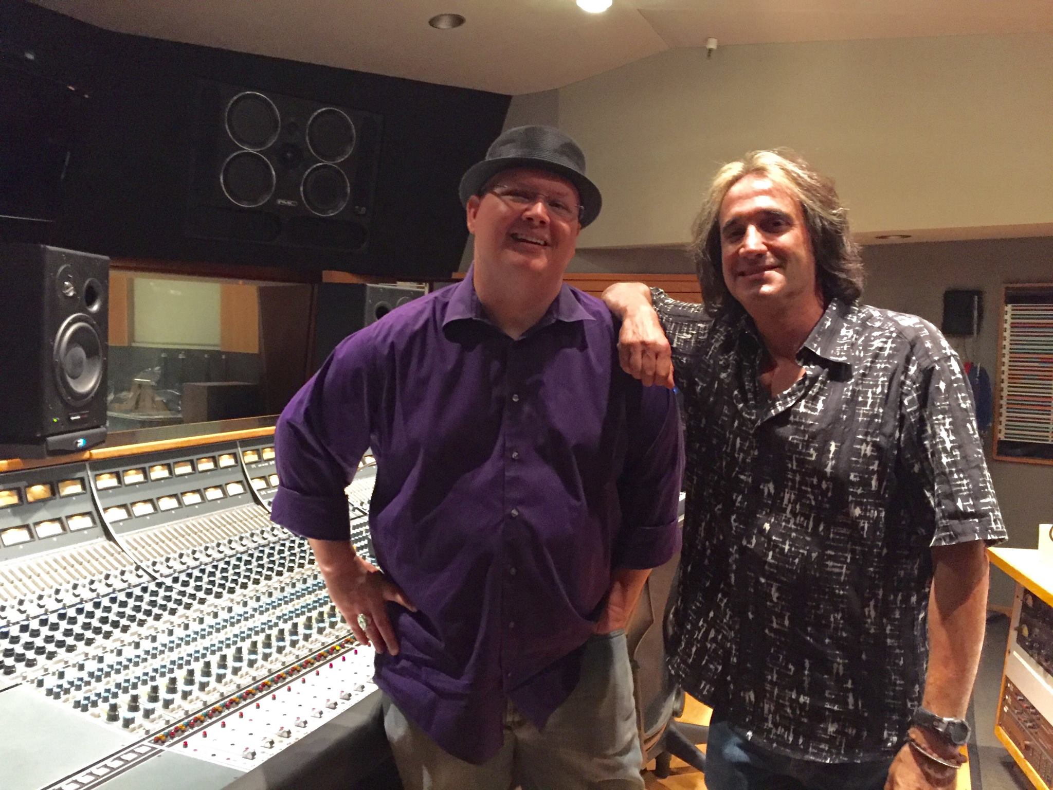 Two men standing in a recording studio, one with a hat and purple shirt, the other with long hair and patterned shirt, smiling.