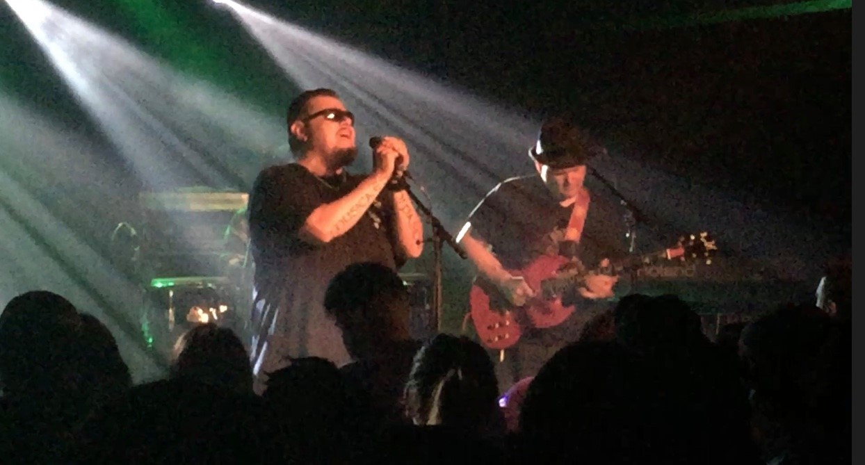 Two musicians perform on stage with bright lights, one singing into a microphone and the other playing guitar, audience in foreground.